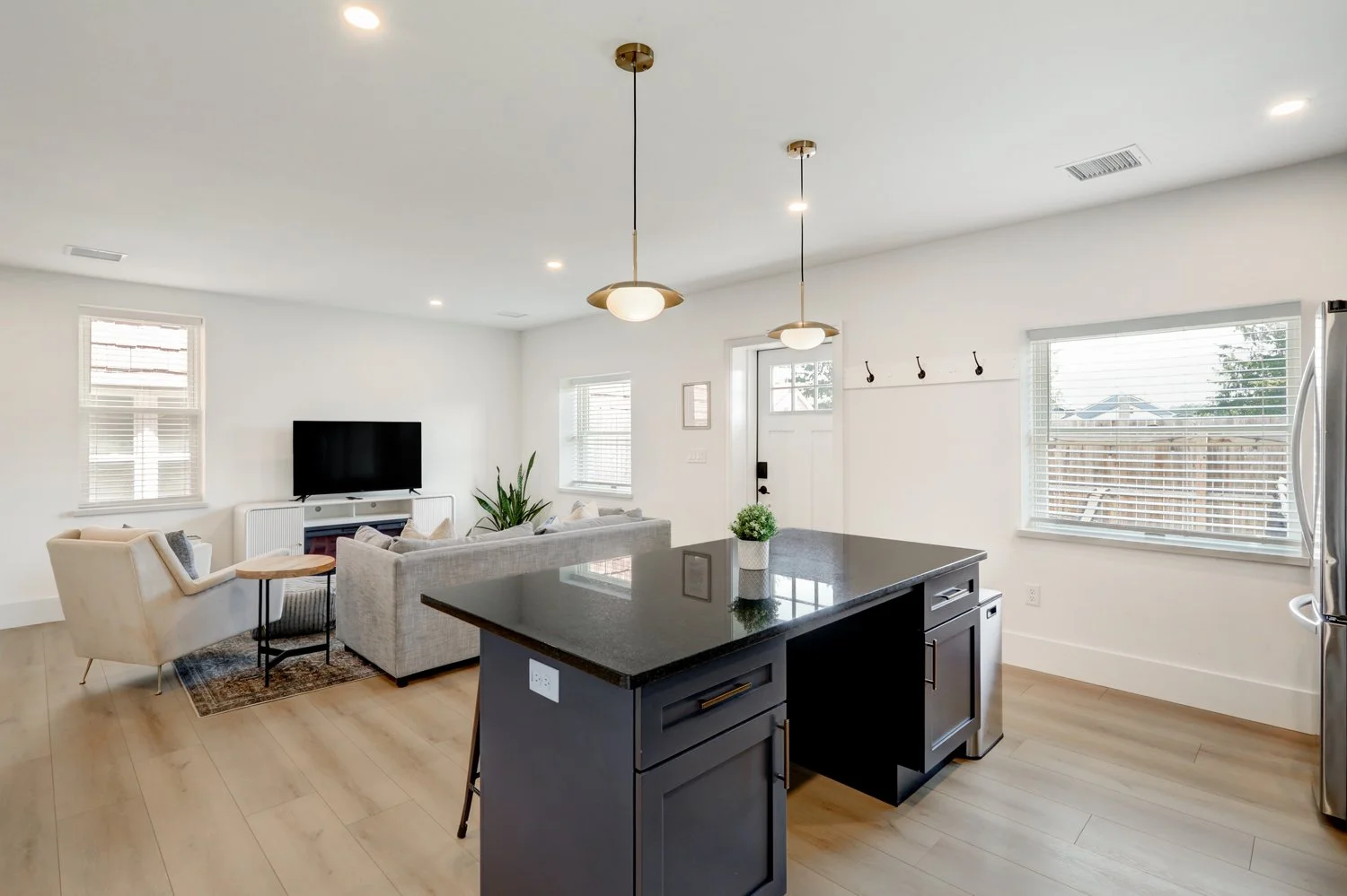 Modern living room and kitchen area with black countertop island, gray sofa, beige armchair with small side table, flat-screen TV on white stand, multiple windows with blinds, ceiling lights, and wooden flooring.