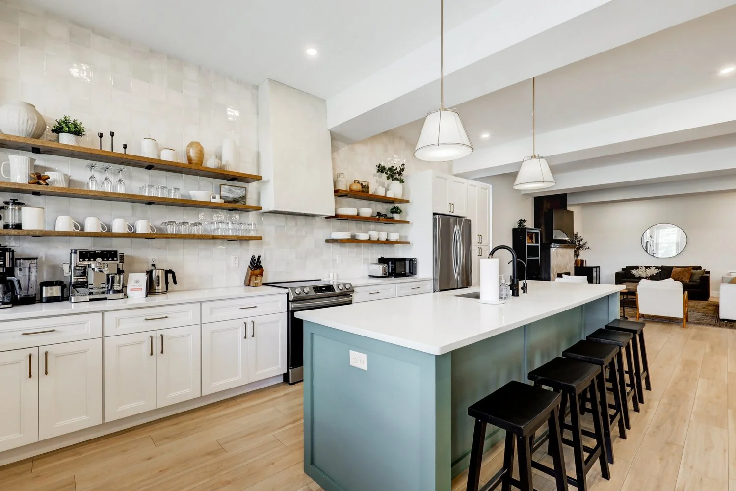 Modern kitchen with white cabinets, a large central island with a teal base, open wooden shelves with dishes and glassware, and living room in the background with seating area.