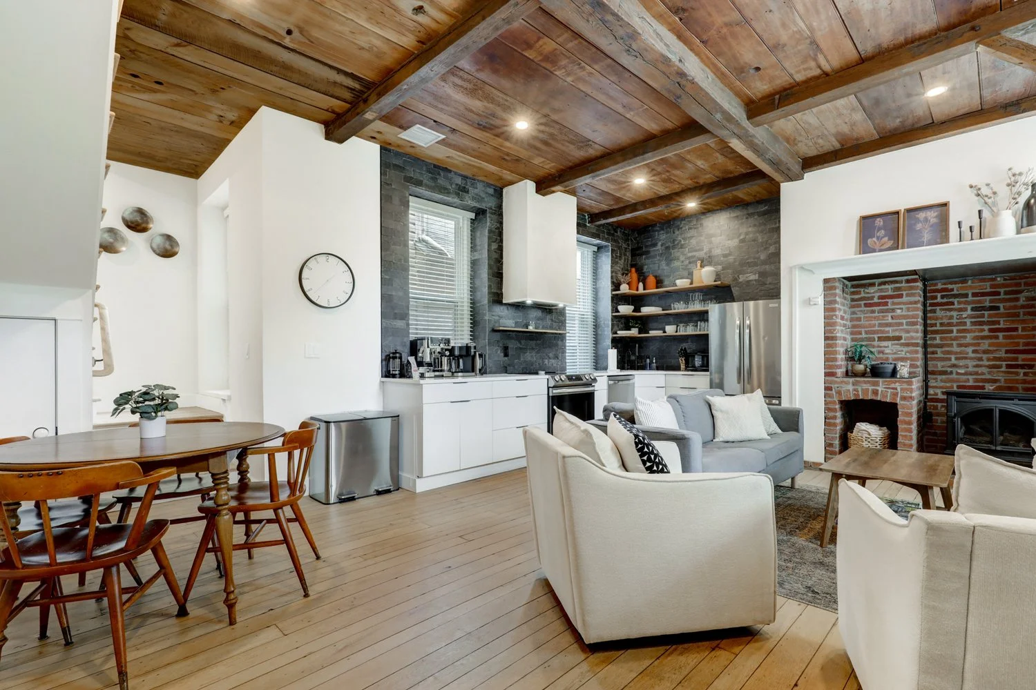 Open-concept living room and kitchen with wooden ceiling, brick fireplace, light-colored furniture, and modern appliances.
