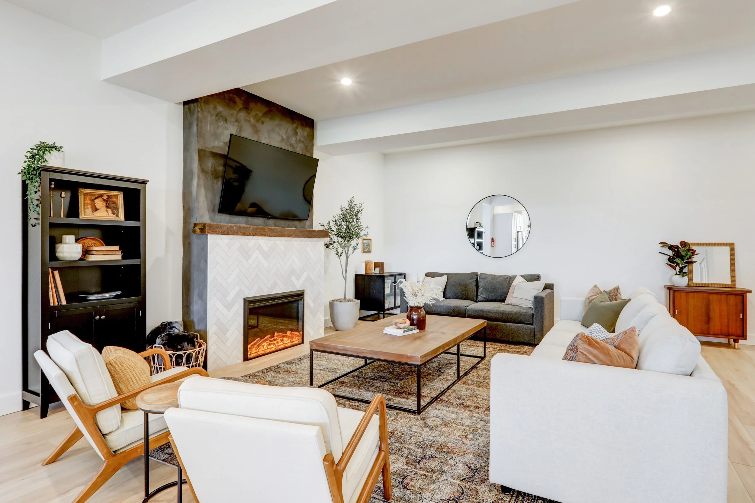 Modern living room with white walls, a fireplace, a flat-screen TV, and various seating including a white chair with wooden frame, a white couch, and dark gray couch. Decor includes plants, framed pictures, a mirror, and a wooden coffee table on a pa