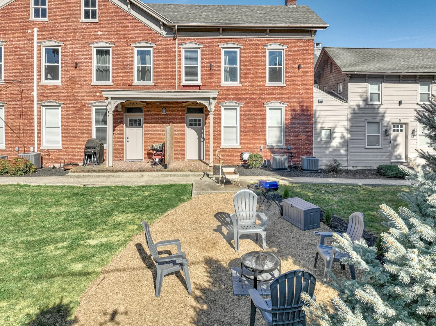 Backyard patio with outdoor furniture including chairs, a fire pit, and a small portable grill. The yard has a combination of grass and gravel, and the back of a red brick residential building with multiple windows and doors. There are two air condit