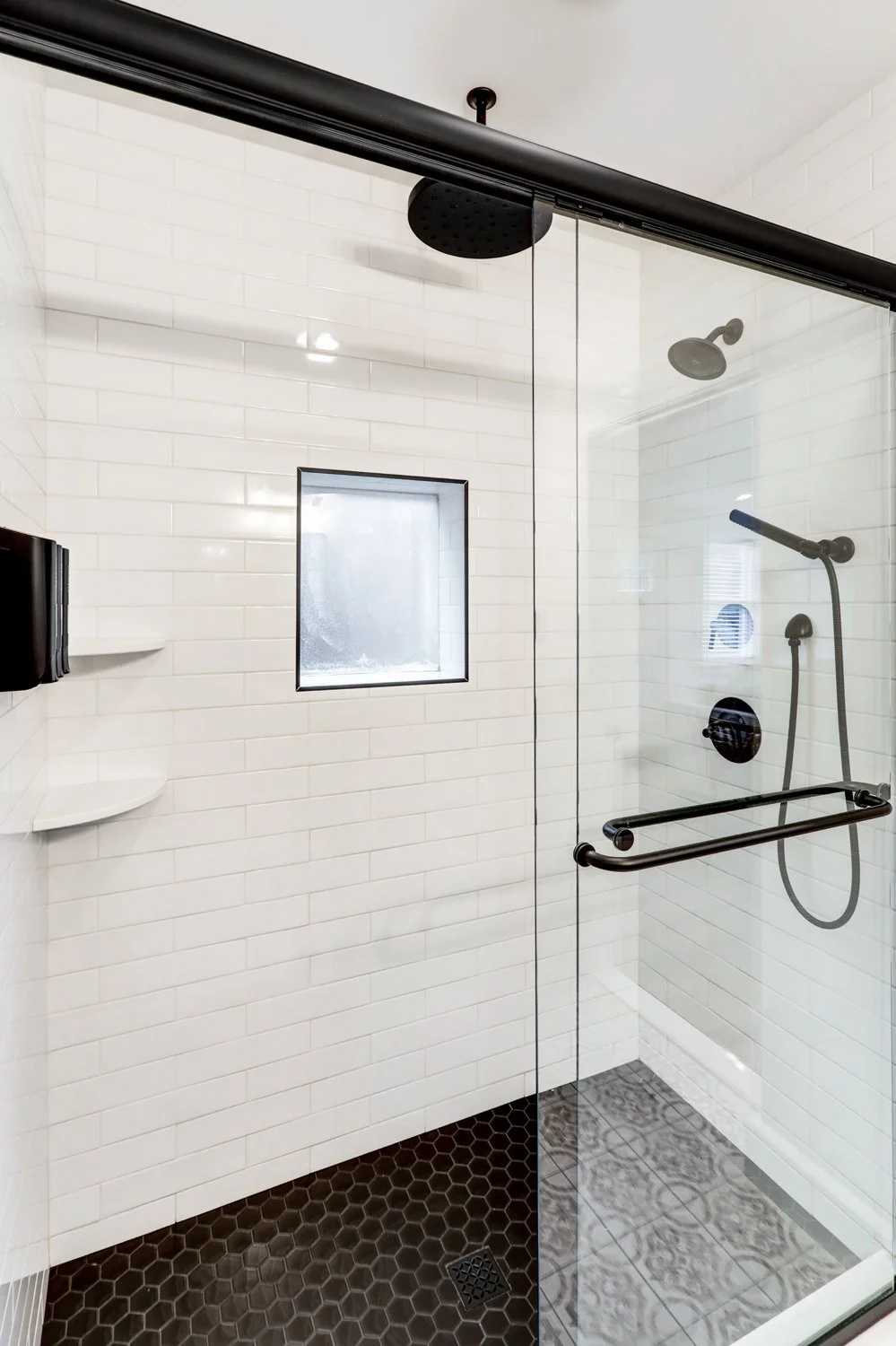A modern walk-in shower with white subway tile walls, black fixtures, a small frosted window, and a glass door. The shower has a rain showerhead and a handheld showerhead, with black accents and a black hexagonal tile floor.