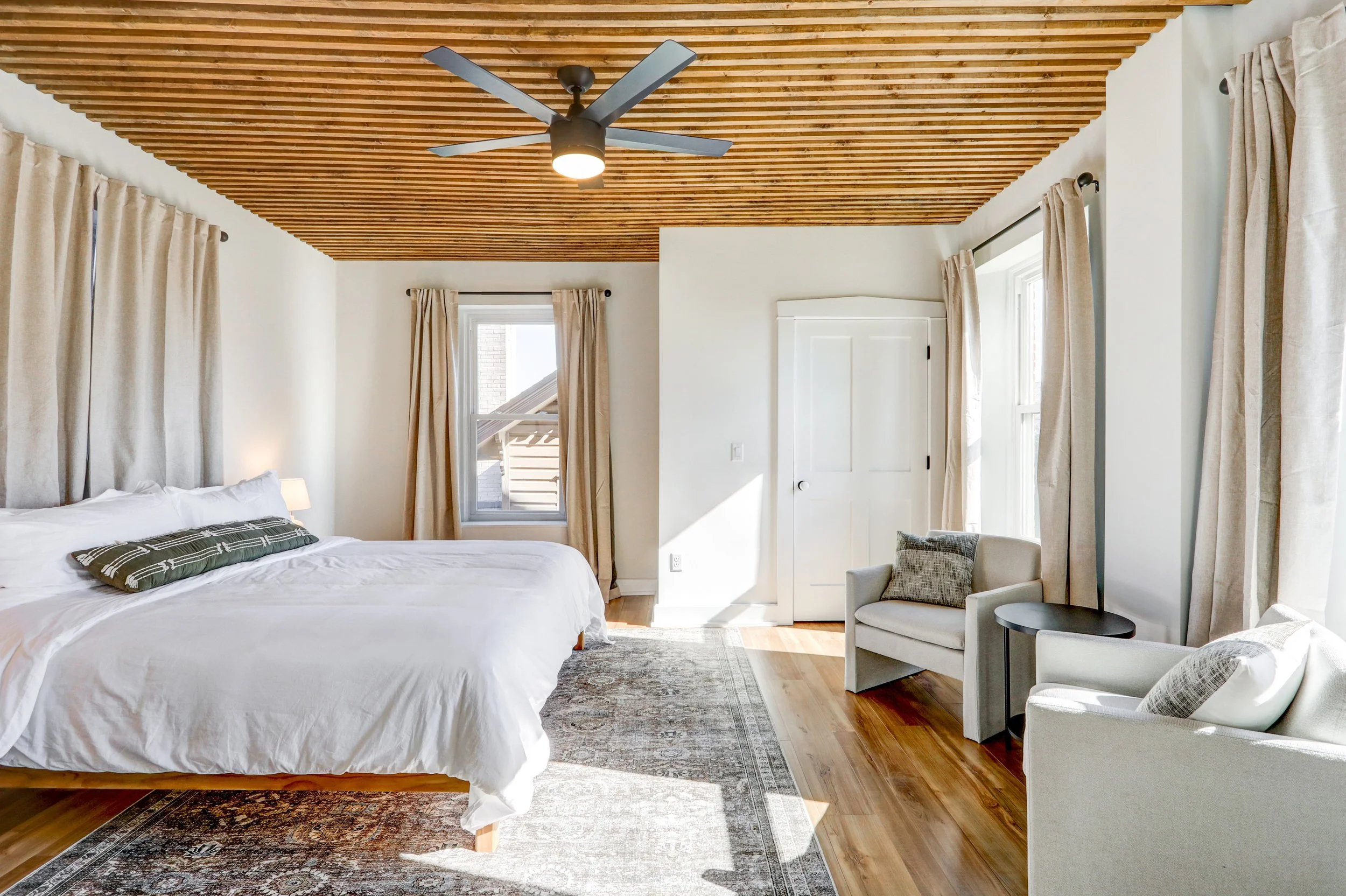 A bedroom with a wood slat ceiling, white walls, and large windows with beige curtains. There is a bed with white bedding and a decorative pillow, a small bedside table with a lamp, a patterned rug on the wooden floor, and a sitting area with a chair
