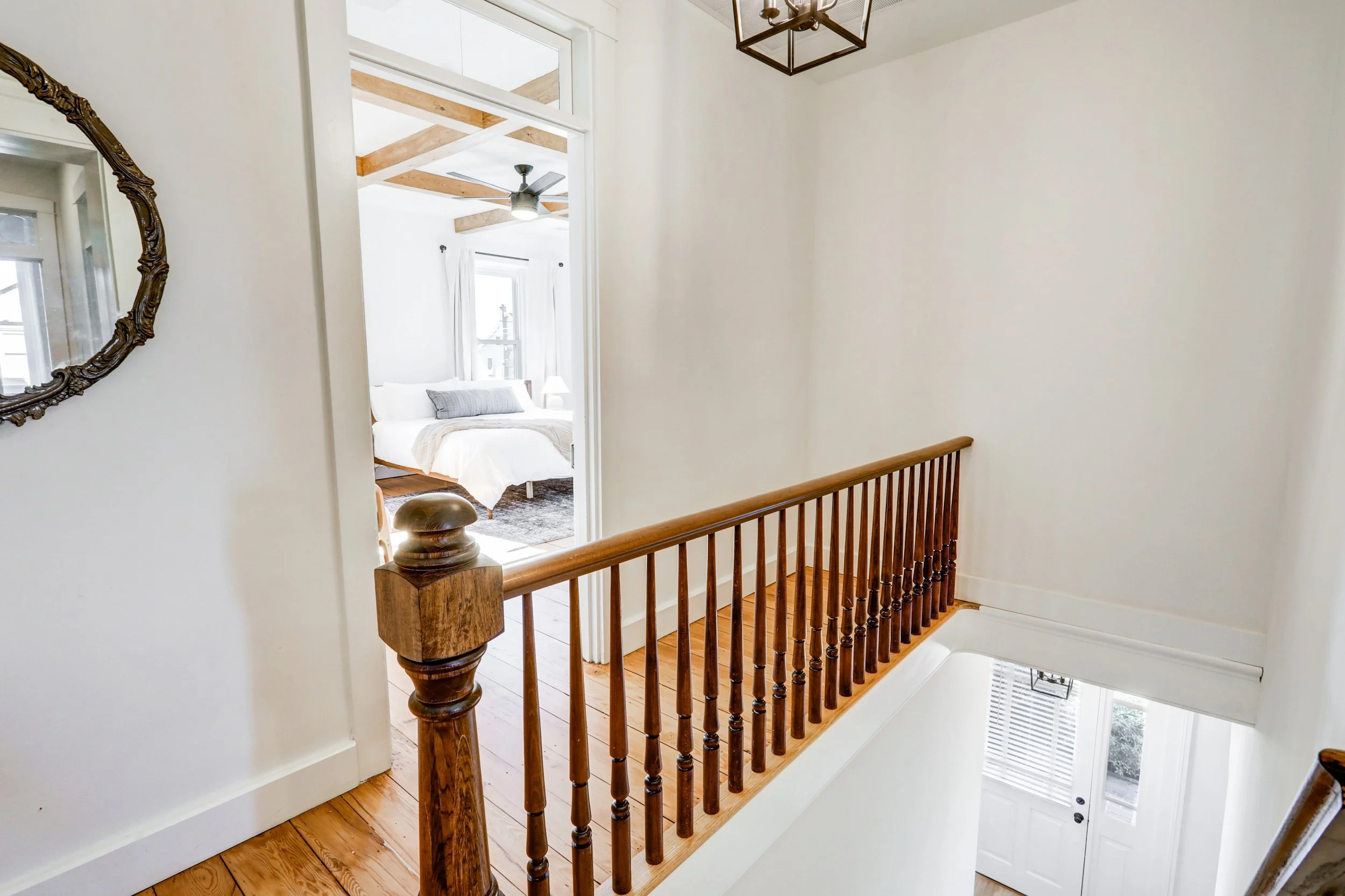 View of a staircase landing with a wooden railing, looking into a bedroom with white walls, a ceiling fan, and a bed with white bedding.