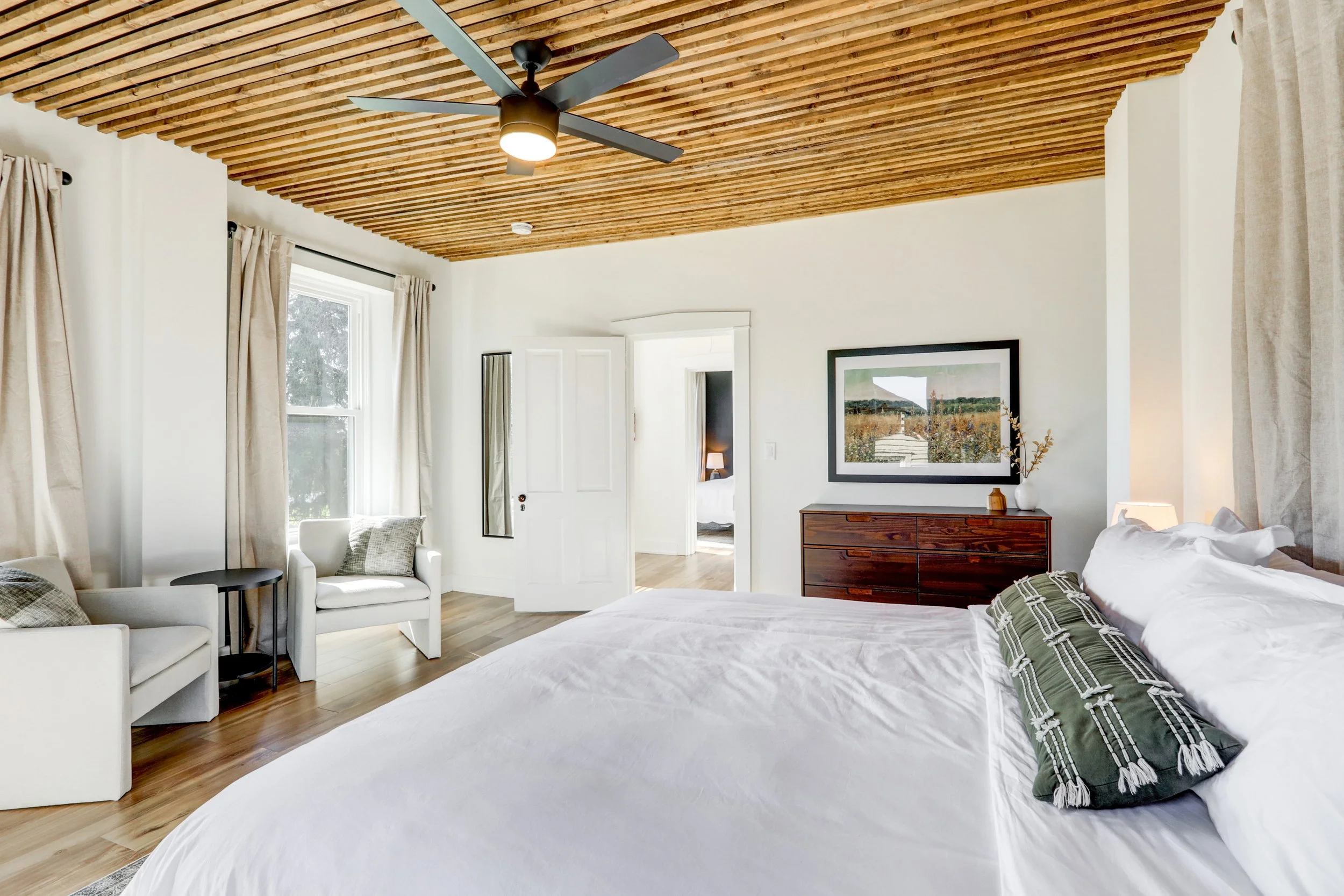 A bedroom with a wooden slatted ceiling, white walls, large windows with beige curtains, a white armchair, a wooden dresser, a framed landscape painting, and a bed with patterned pillows.