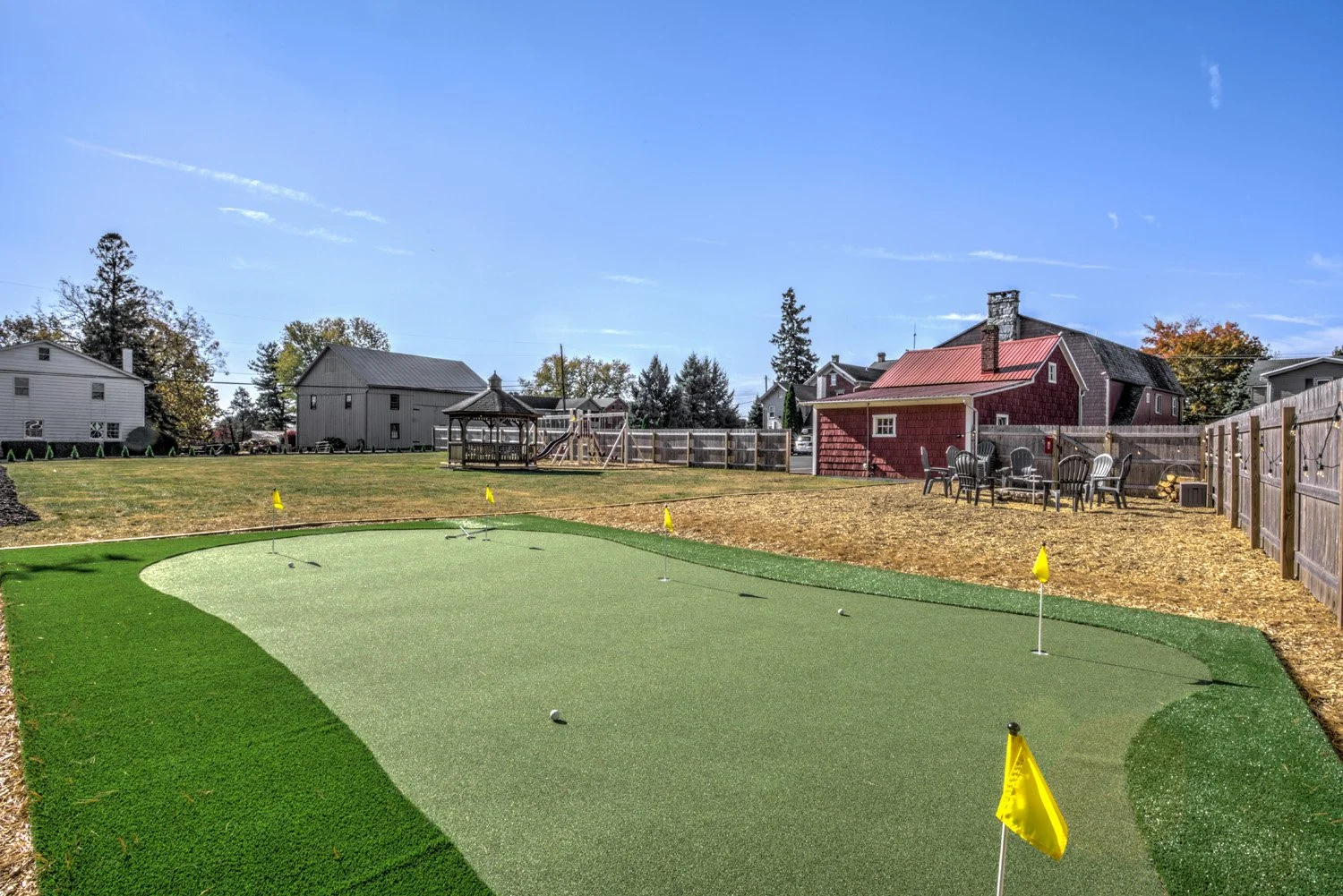 A backyard with a mini golf putting green, yellow flagsticks, and a golf ball. The yard has a fenced area with outdoor furniture and a red barn-style house in the background. There is a playset with swings and a slide on the far side of the yard unde