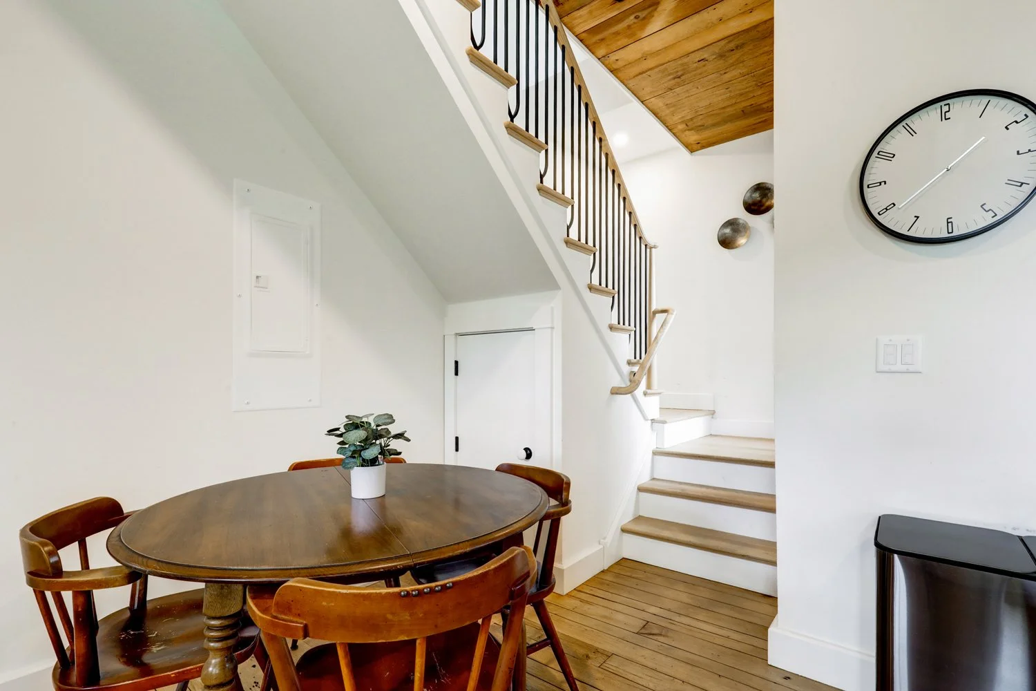 Interior view of a home with a wooden staircase, round dining table with four chairs, a potted plant, a wall clock, and a small black cabinet.
