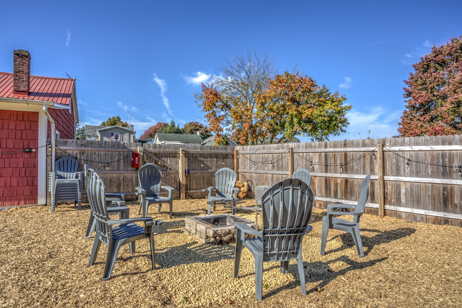 Backyard with wooden fence, outdoor chairs arranged in a circle around a fire pit, and trees with fall foliage under a blue sky.