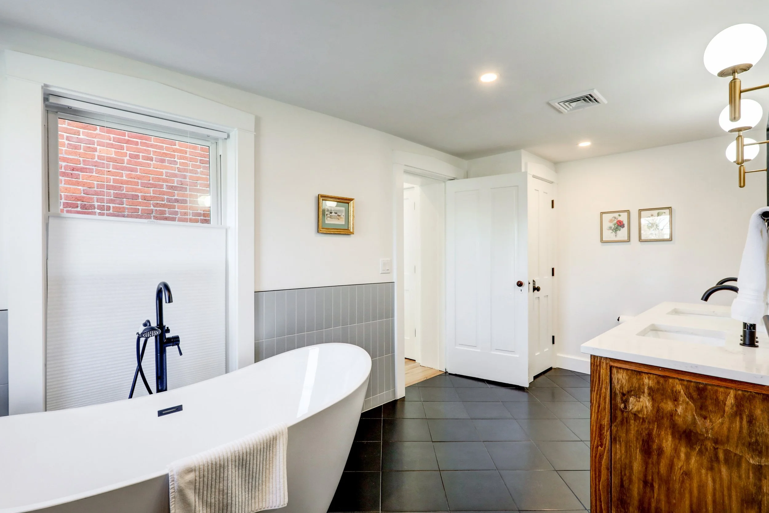 A bathroom with a freestanding bathtub, a black faucet, a window showing a red brick wall outside, a wooden vanity with a white countertop and double sinks, wall art of flowers, and modern ceiling lights.