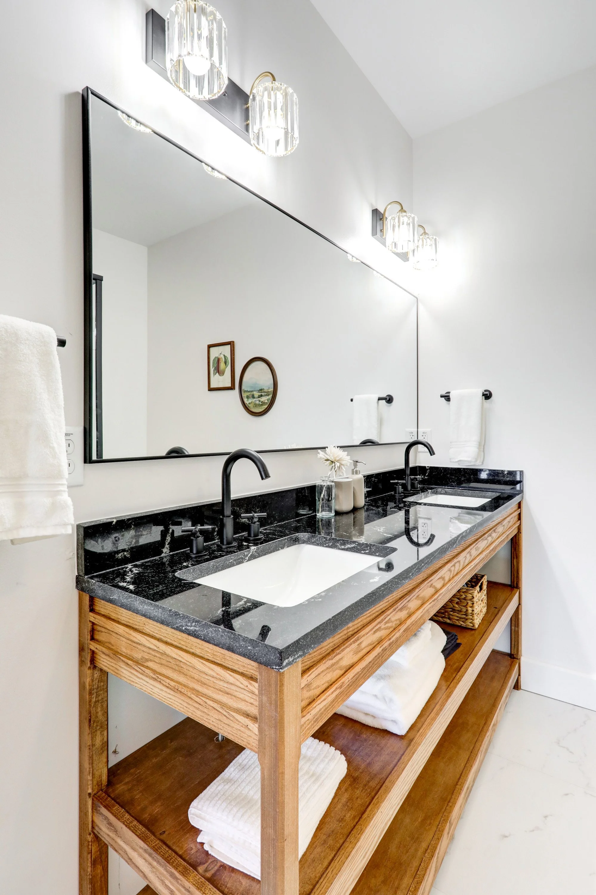 Modern bathroom with a large mirror, black sinks, gold and glass wall sconces, a wooden vanity with open shelving, towels, and decorative items.