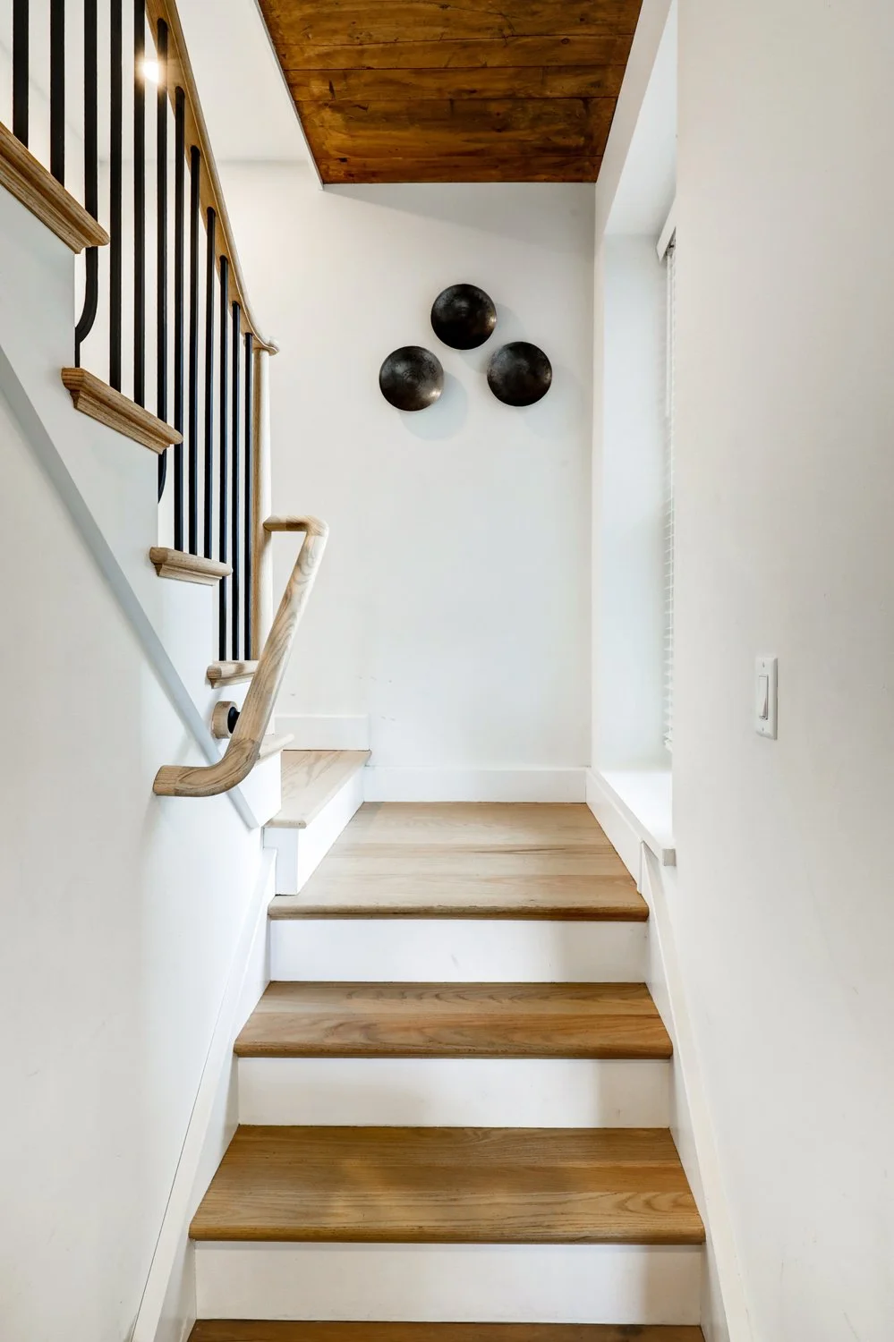 Interior view of a staircase with white walls, wooden steps, a wooden handrail, and a black metal railing on the left side. Three black decorative wall art pieces are on the wall at the top of the stairs, and a window with blinds is on the right side