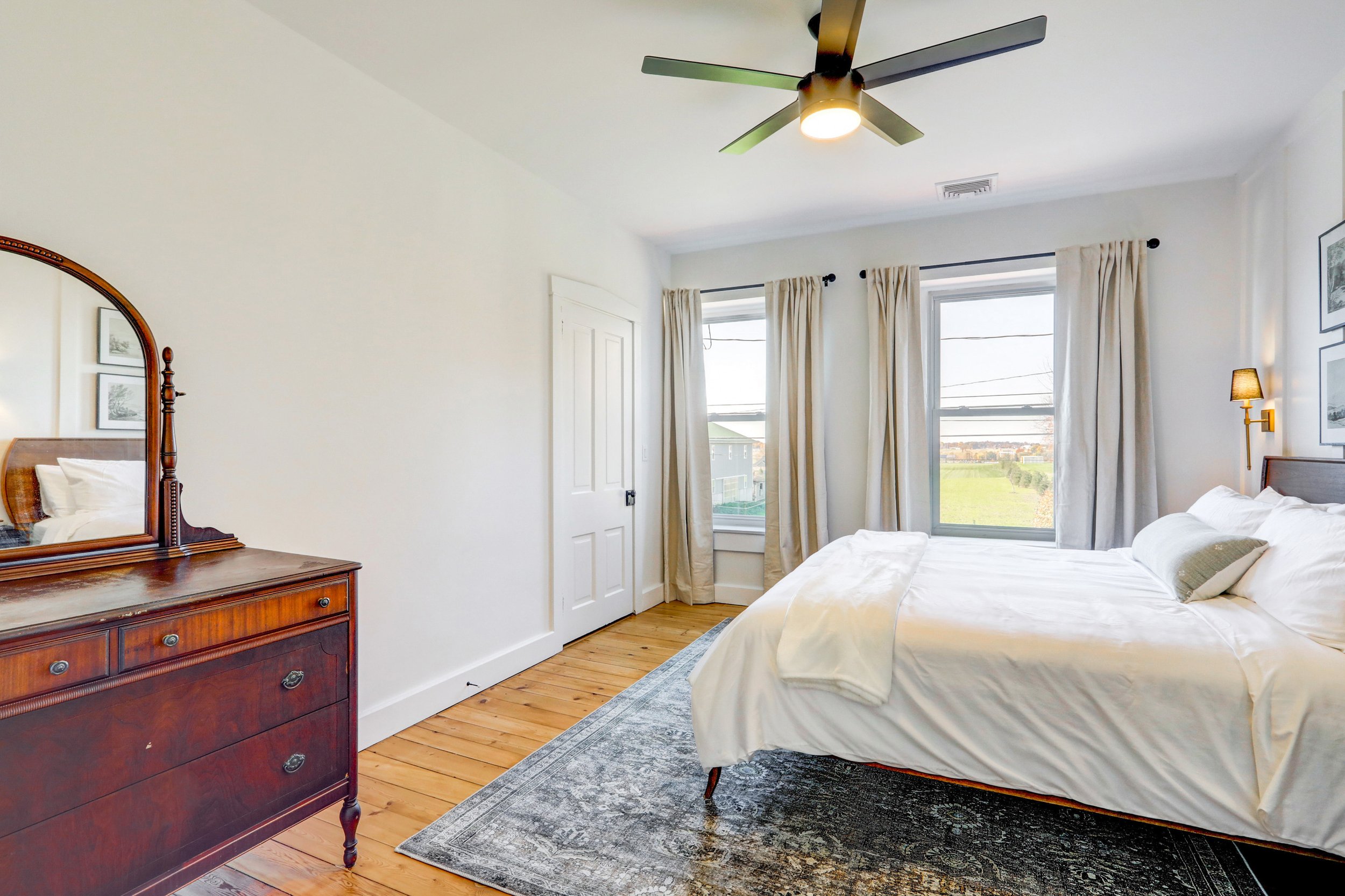 A bedroom with white walls, a wooden dresser with a mirror, a bed with white bedding, windows with cream curtains, a ceiling fan with lights, and framed pictures on the wall.