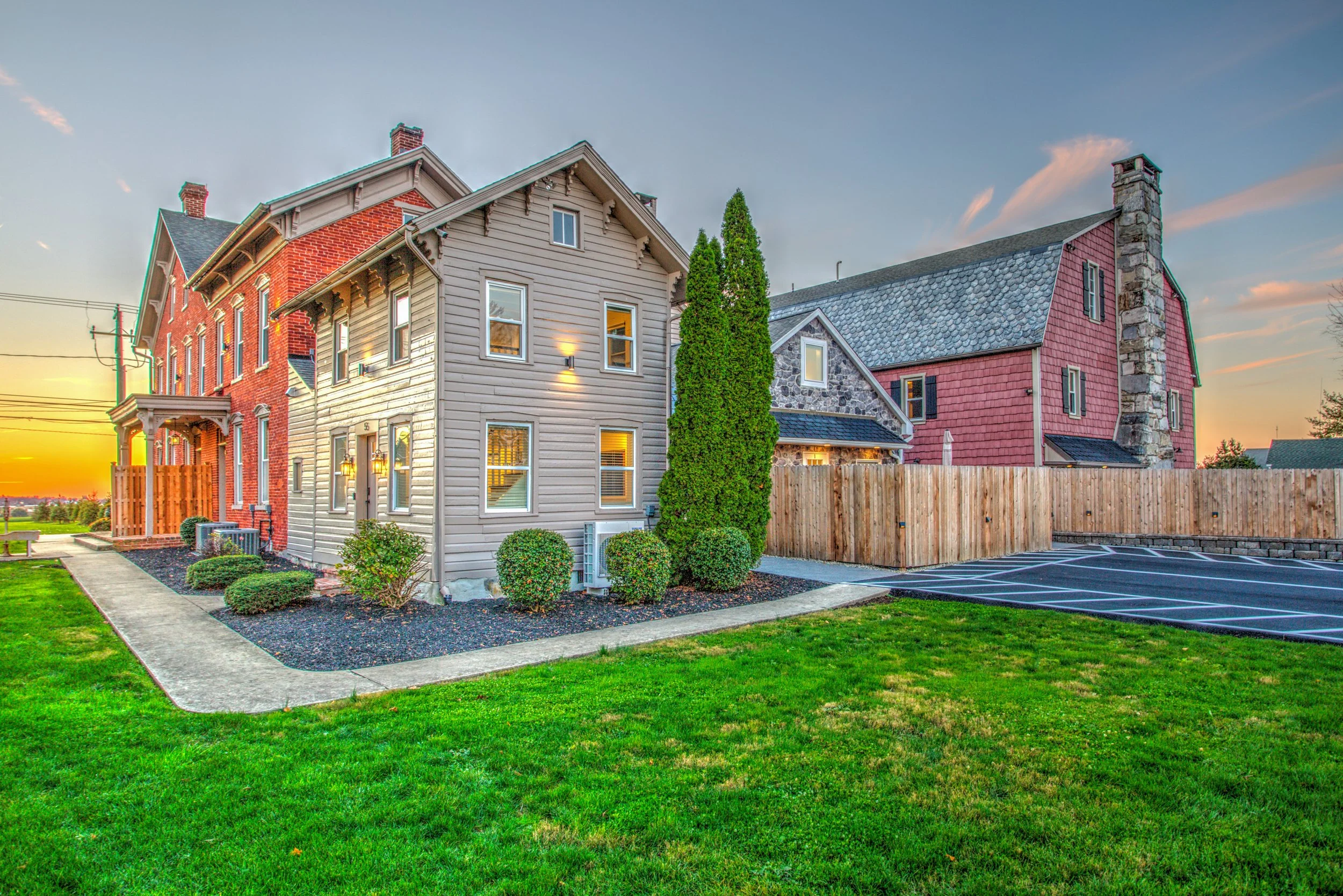 Side view of a residential building with varied architectural styles, including red brick, beige siding, stone, and red shingles, lawn, and parking area, during sunset.