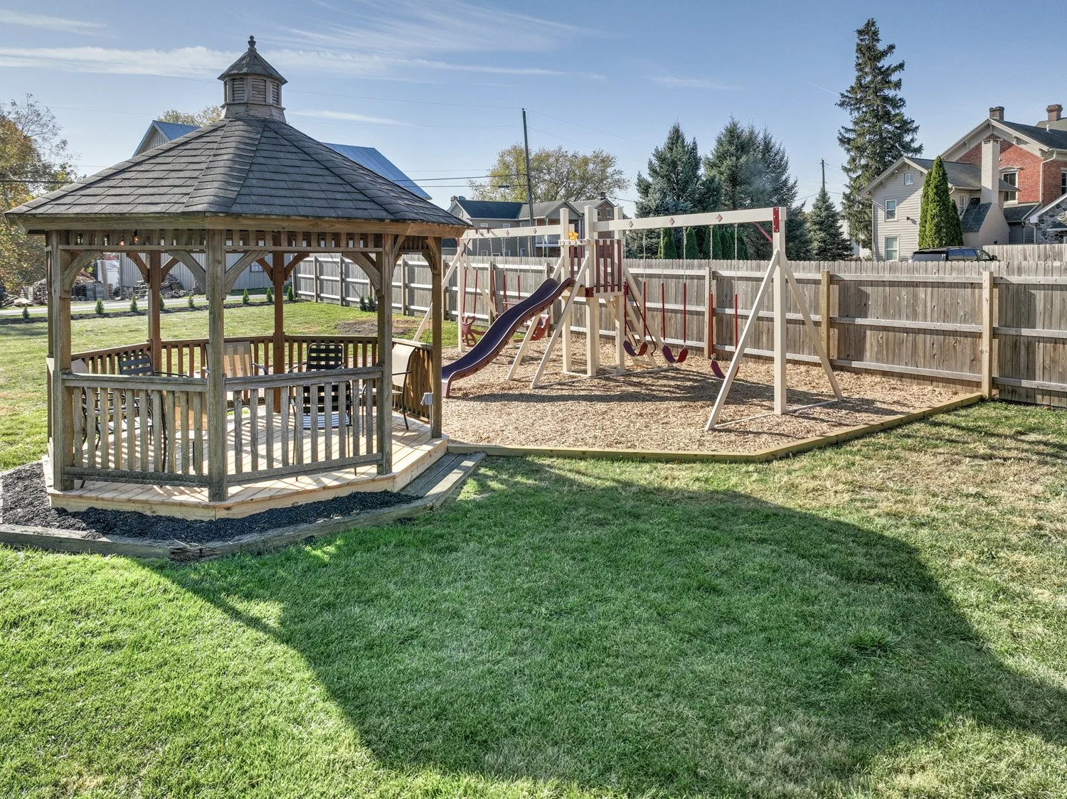 Residential backyard with a wooden gazebo, a swing set, and a fence. The yard has green grass and neighboring houses and trees are visible.
