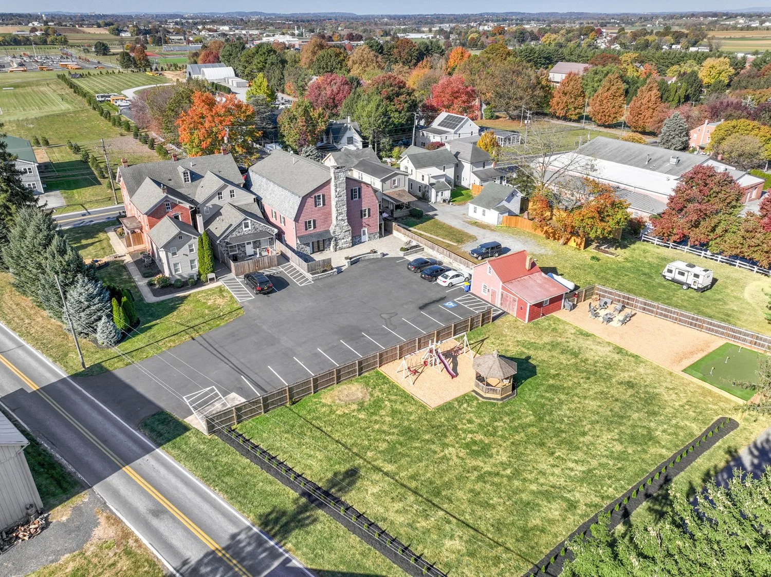 Aerial view of a residential area with a mix of houses and a large fenced backyard featuring a playground with swings and a gazebo, parking lot, and a variety of colorful trees around.