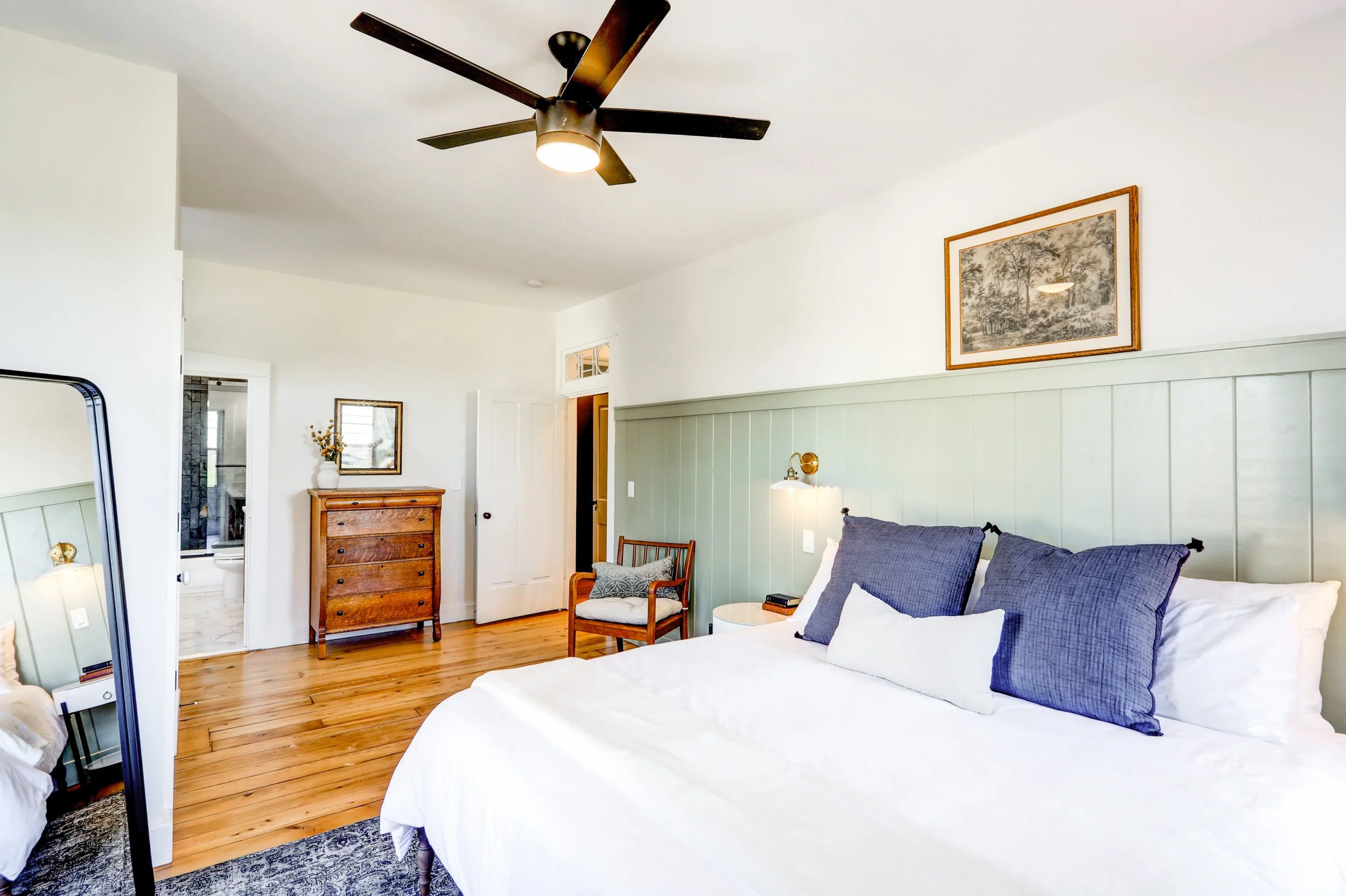 A bedroom with a queen-sized bed featuring navy blue pillows, a wooden dresser, a small wooden chair, and artwork on the white wall. There is a ceiling fan and a window.