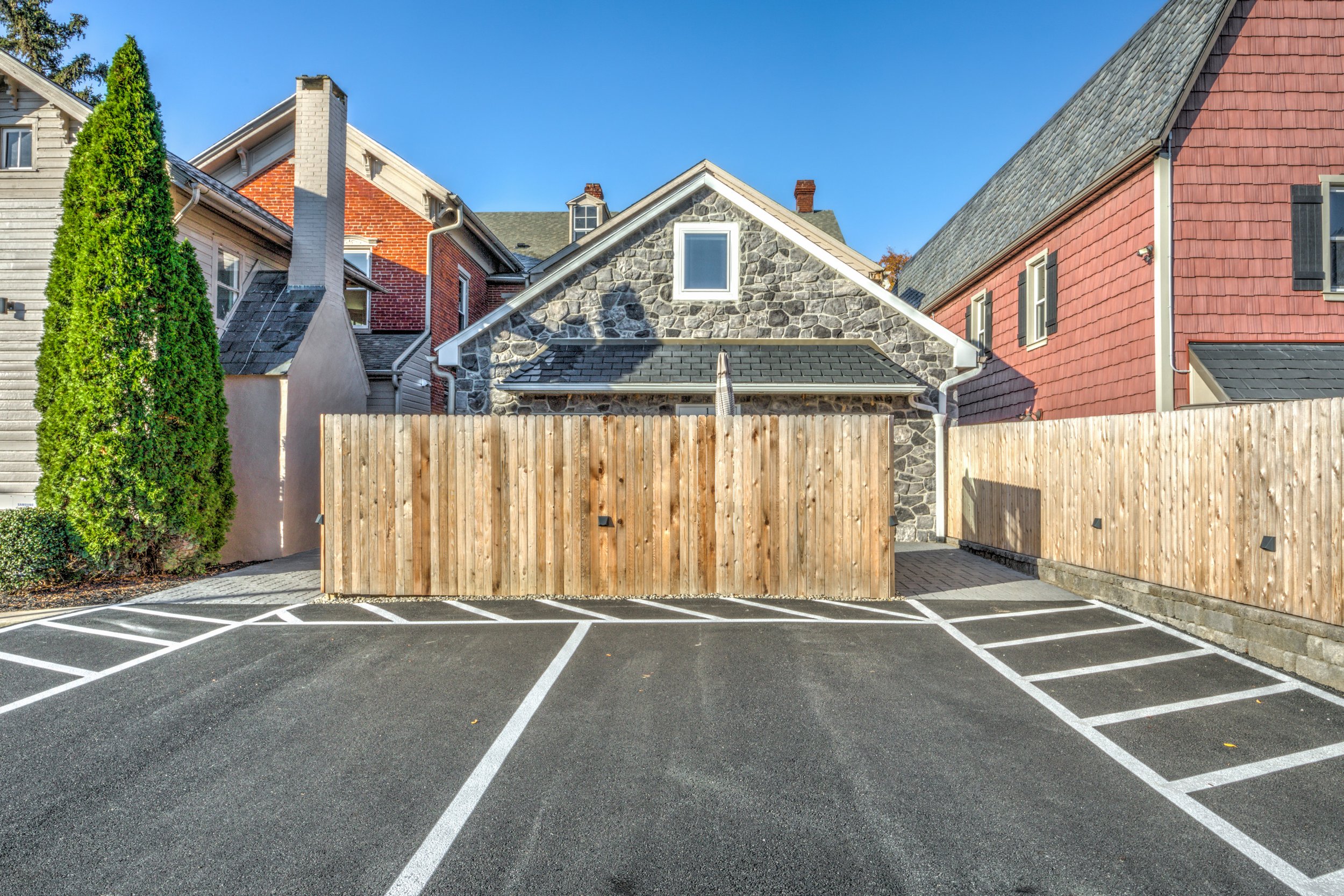 A parking lot with white-painted parking line markings in front of a row of houses with varied architectural styles and materials, including a stone house, brick houses, and a house with light-colored siding, separated by a tall wooden fence.