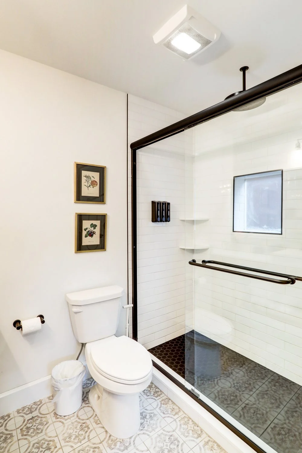 Bathroom with a toilet, trash can, framed botanical prints, and a walk-in shower with white subway tiles, black trim, and a window.