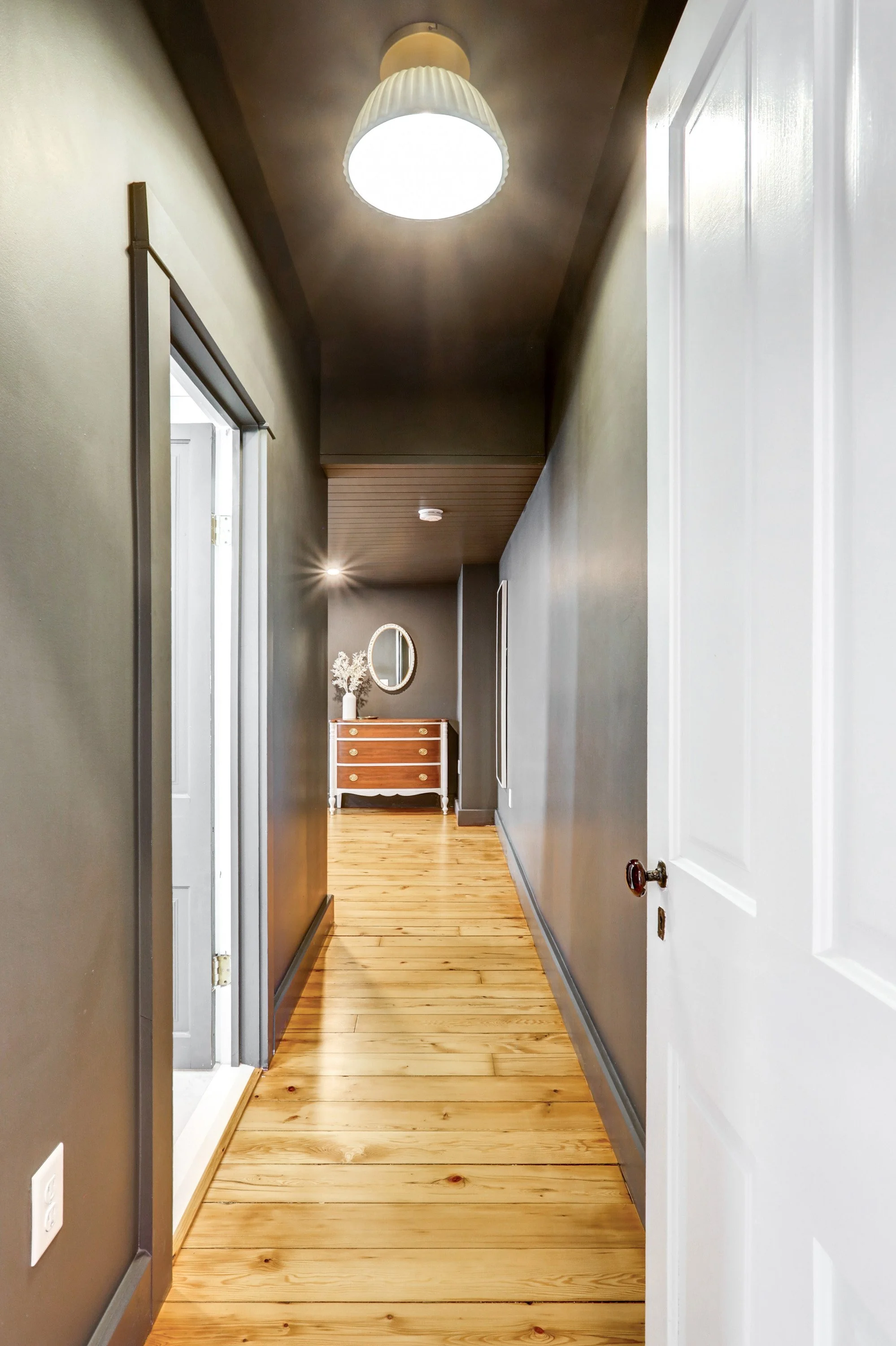 A hallway with light wood flooring, gray and white walls, a ceiling light fixture, and a dresser with a mirror at the end of the hall.