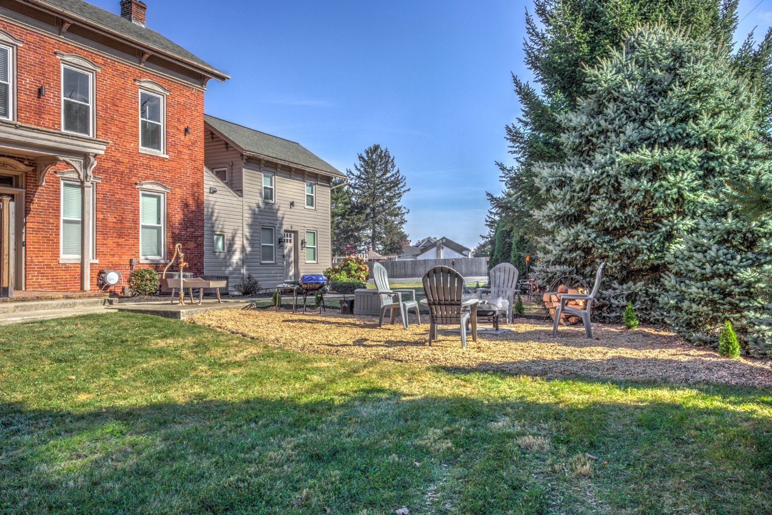 Backyard with outdoor seating area, Adirondack chairs, a small firepit, large pine trees, and a brick and gray house in the background on a sunny day.
