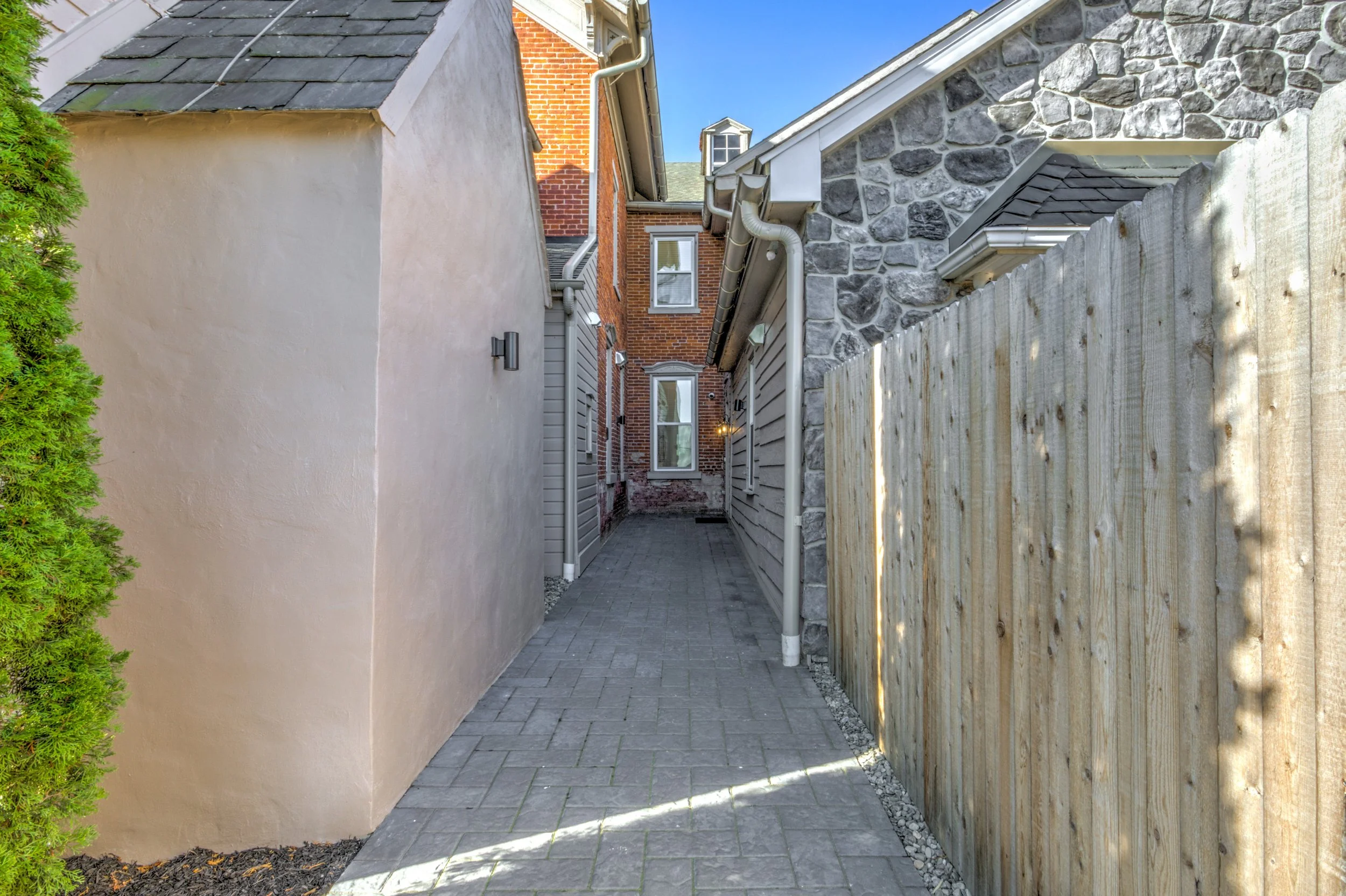 A narrow paved alleyway between a beige stucco house and a wooden fence, with a brick house in the background, under a clear blue sky.