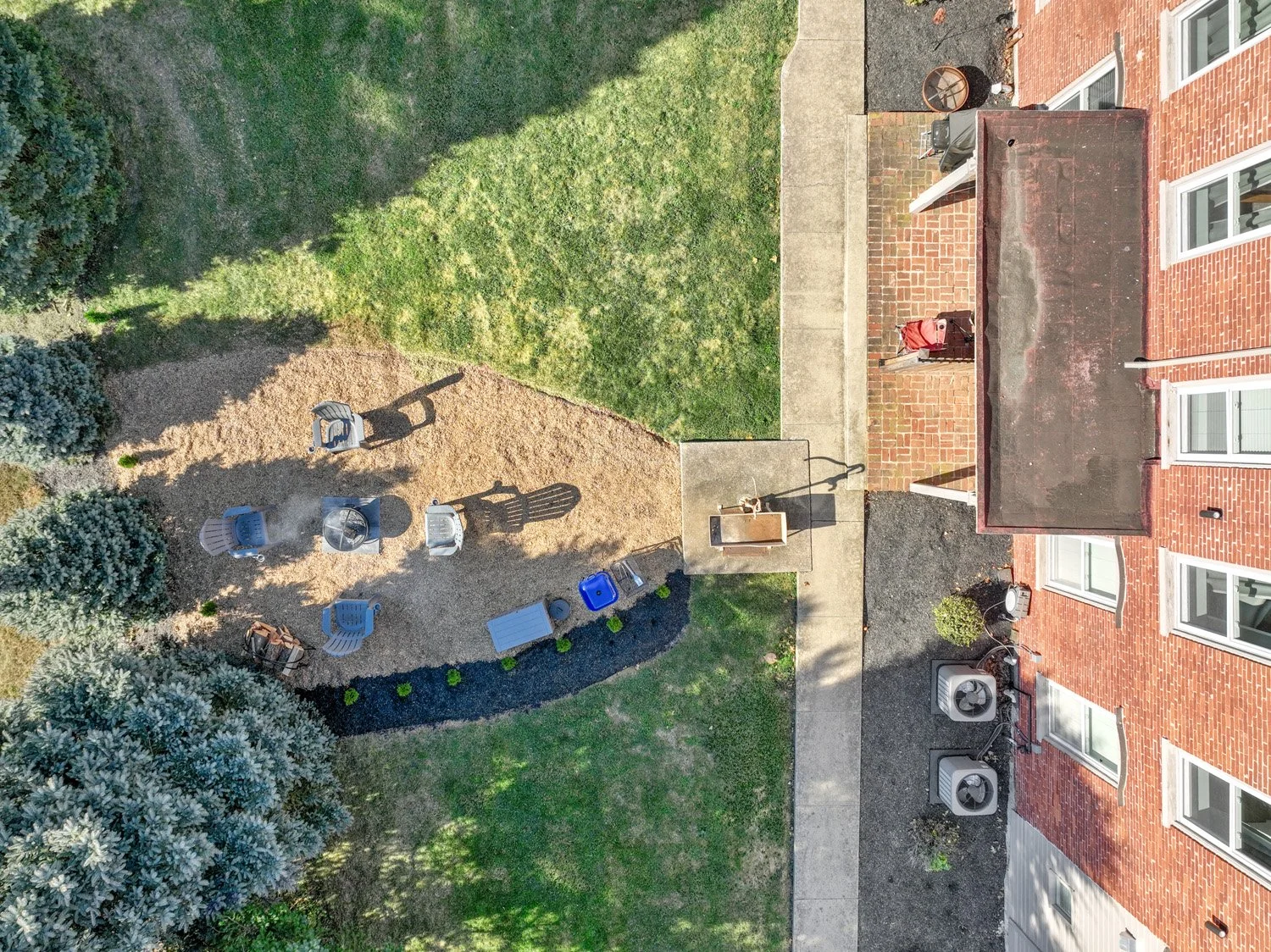 Bird's-eye view of an apartment backyard with a green lawn, a sandy area with outdoor chairs, and a concrete pathway. The brick building has several windows, air conditioning units, and a small covered patio area.