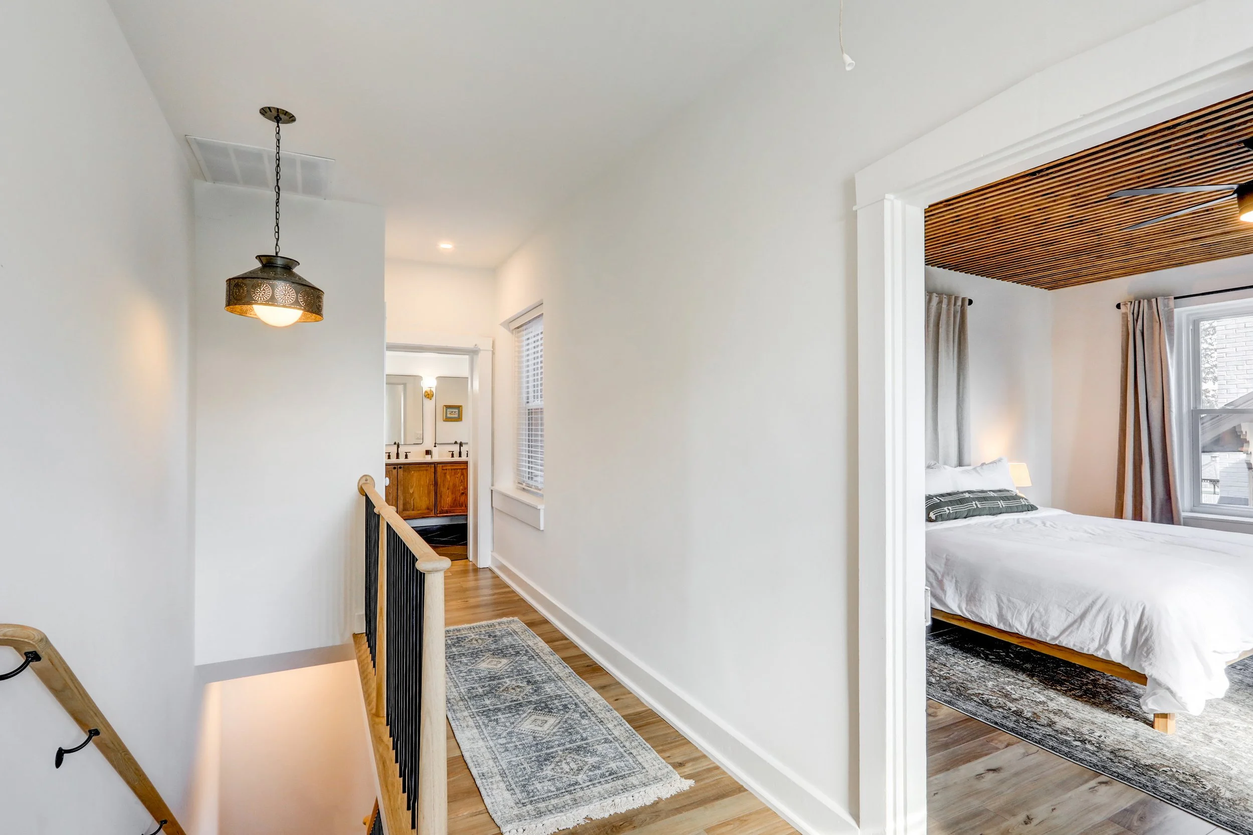 Interior view of a home hallway with white walls, wooden flooring, and a staircase. A hanging light fixture with a decorative pattern is above the stairs. The hallway leads to a bedroom with a bed, curtains, and windows visible. There is another room