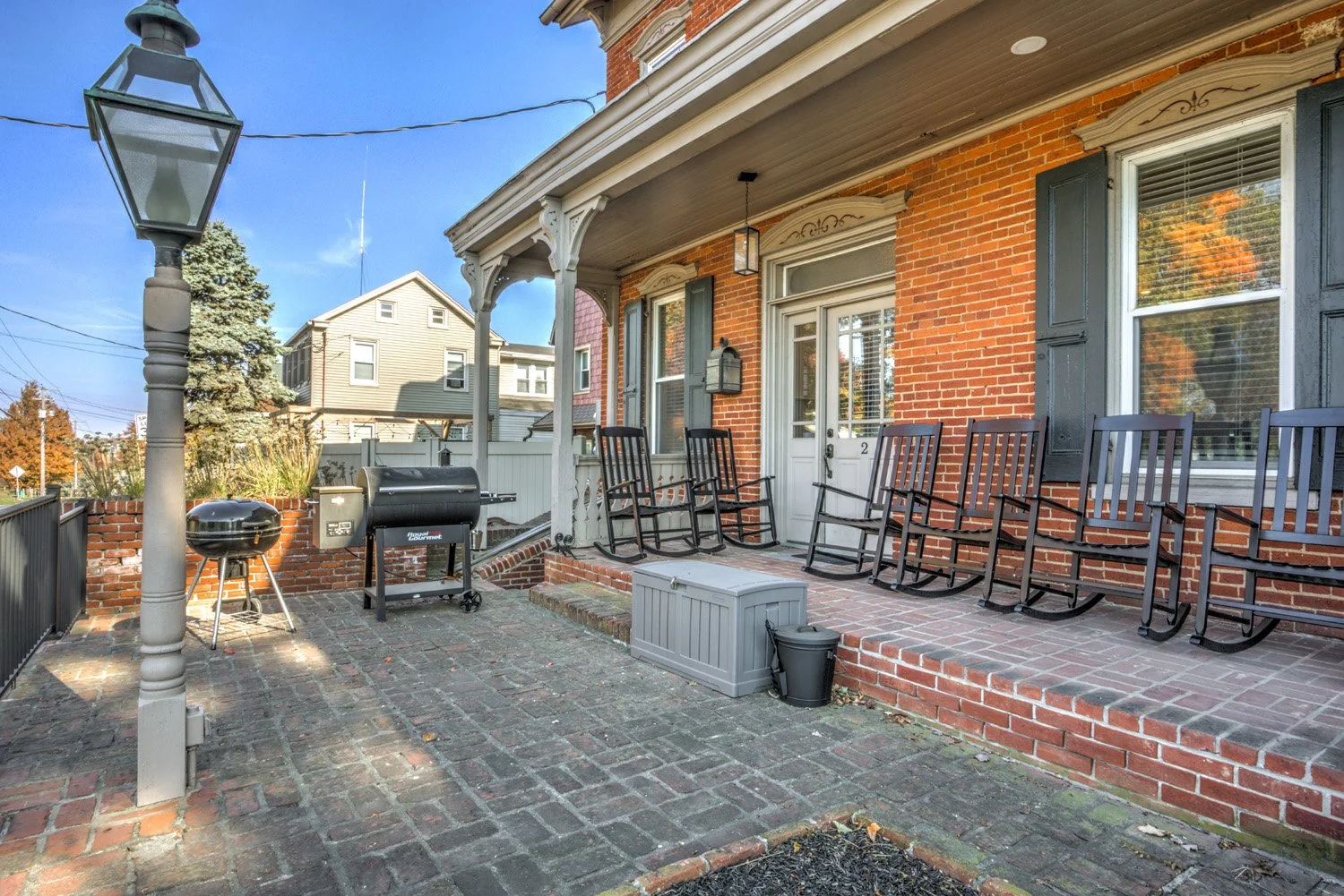 Brick porch with black rocking chairs, a white door, a black lantern, and a small gray storage bench. There are two grills and a string light, with a tree and houses in the background under a blue sky.