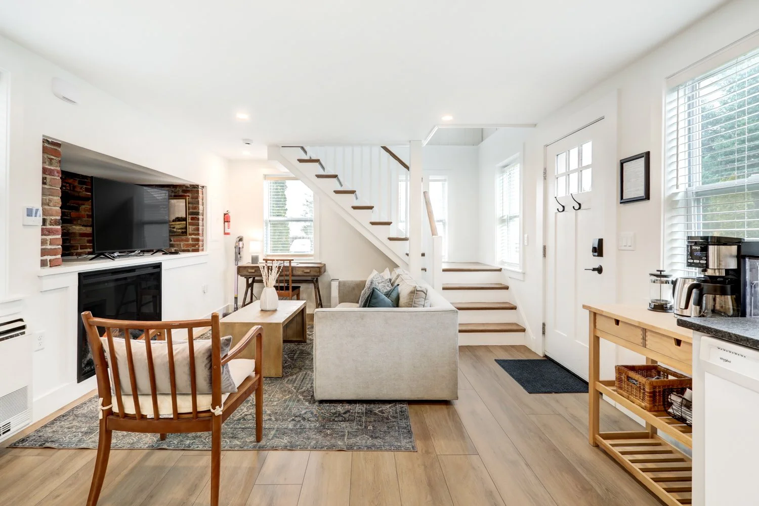 Bright living room with white walls, wooden flooring, sofa, armchair, coffee table, staircase, window blinds, TV, fireplace, and kitchen area with coffee maker and toaster.