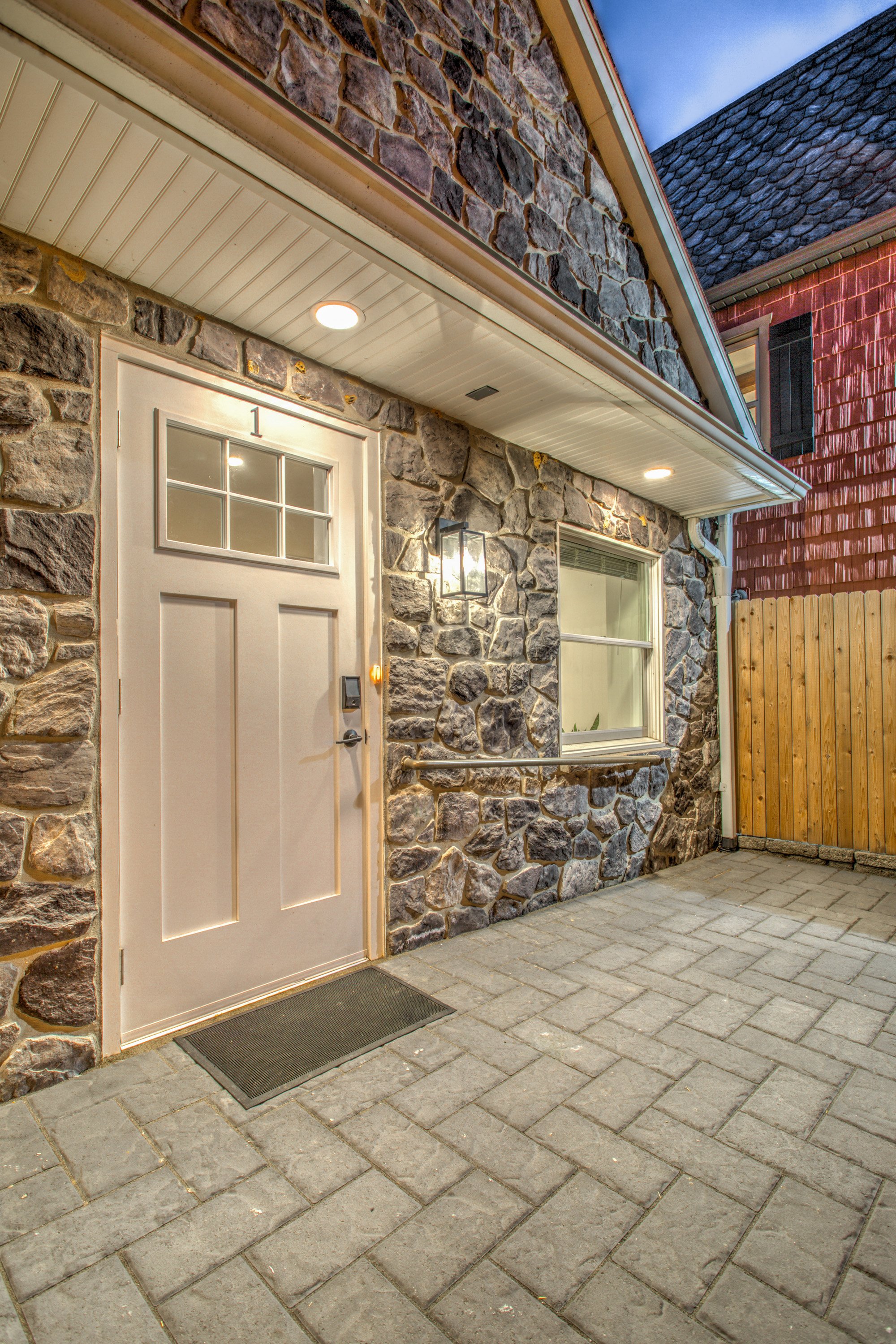 Exterior of a house with a stone facade, a white door with the number 1, a window, and a wooden fence nearby. The house has a sloped roof with shingles and is illuminated by outdoor lights.