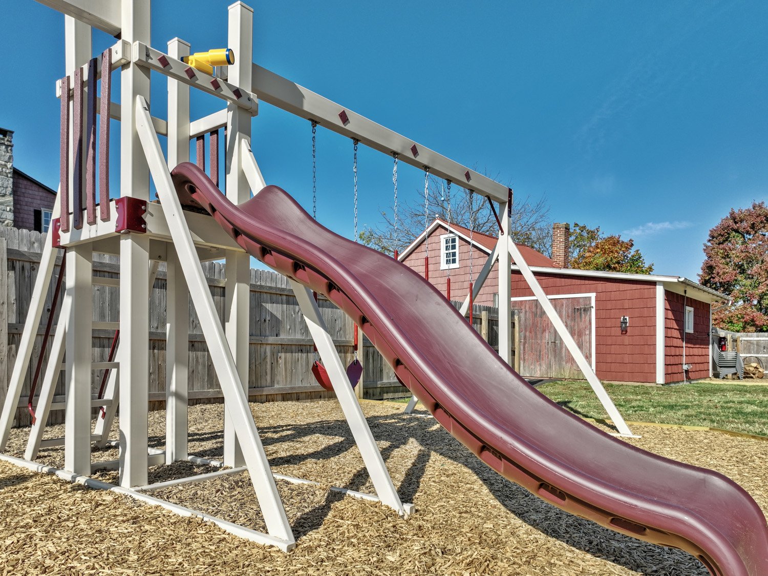 A backyard playground with a red slide, a swing set, and a wooden fence. There is a red shed and some trees with fall foliage in the background, under a clear blue sky.