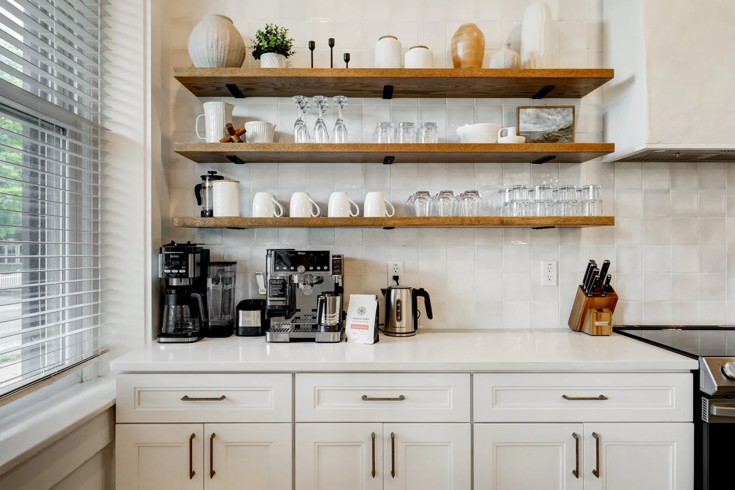 Clean kitchen countertop with coffee machine, espresso maker, electric kettle, and knife block. Open wooden shelves above hold various cups, glasses, and decorative vases. White cabinets below and natural light from window with blinds.
