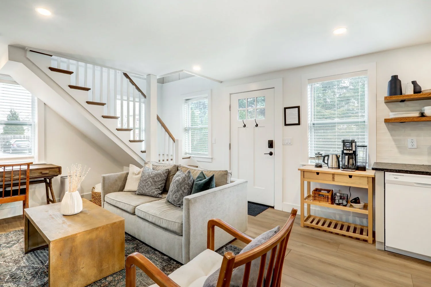 Living room with sofa, wooden coffee table, and stairs near the front door, kitchen area with open shelves, coffee maker, and windows with blinds.
