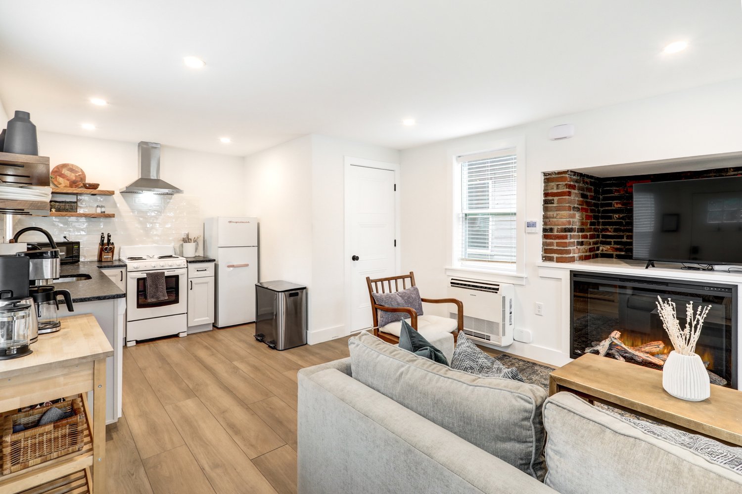 Living room and kitchen with white walls, wood flooring, brick fireplace, sofa with pillows, chair, TV, refrigerator, stove, countertops, open shelves, and various kitchen appliances.