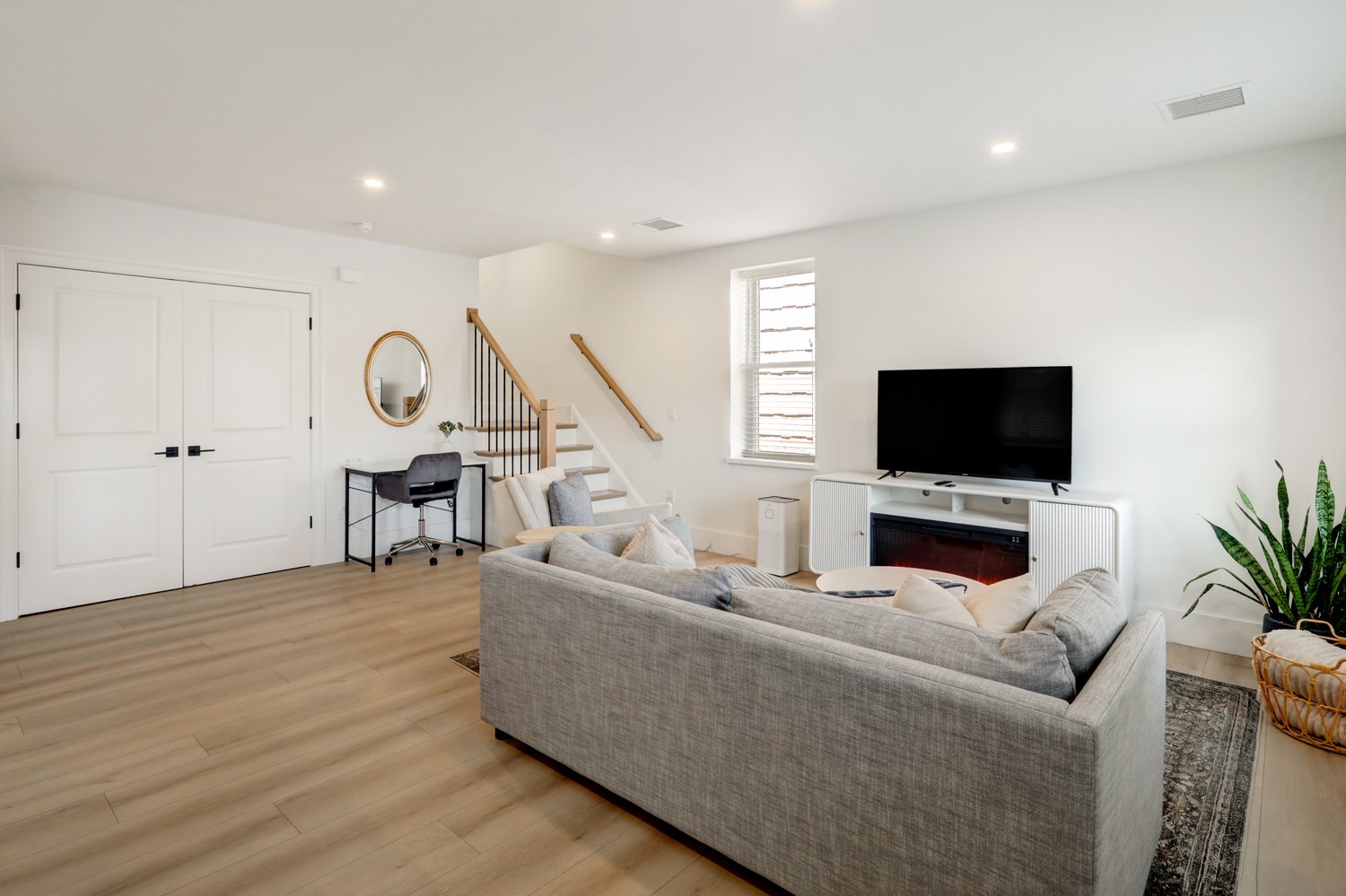 Modern living room with white walls and wooden flooring, featuring a gray sofa, a flat-screen TV on a white media console, a small desk with a chair near a staircase, a window with blinds, and potted plants.
