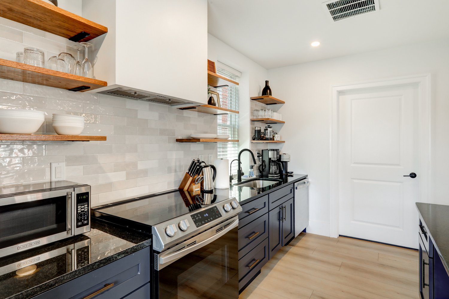 Modern kitchen with black countertops, blue cabinets, open wooden shelves, white tiled backsplash, stainless steel appliances, and a window with blinds.