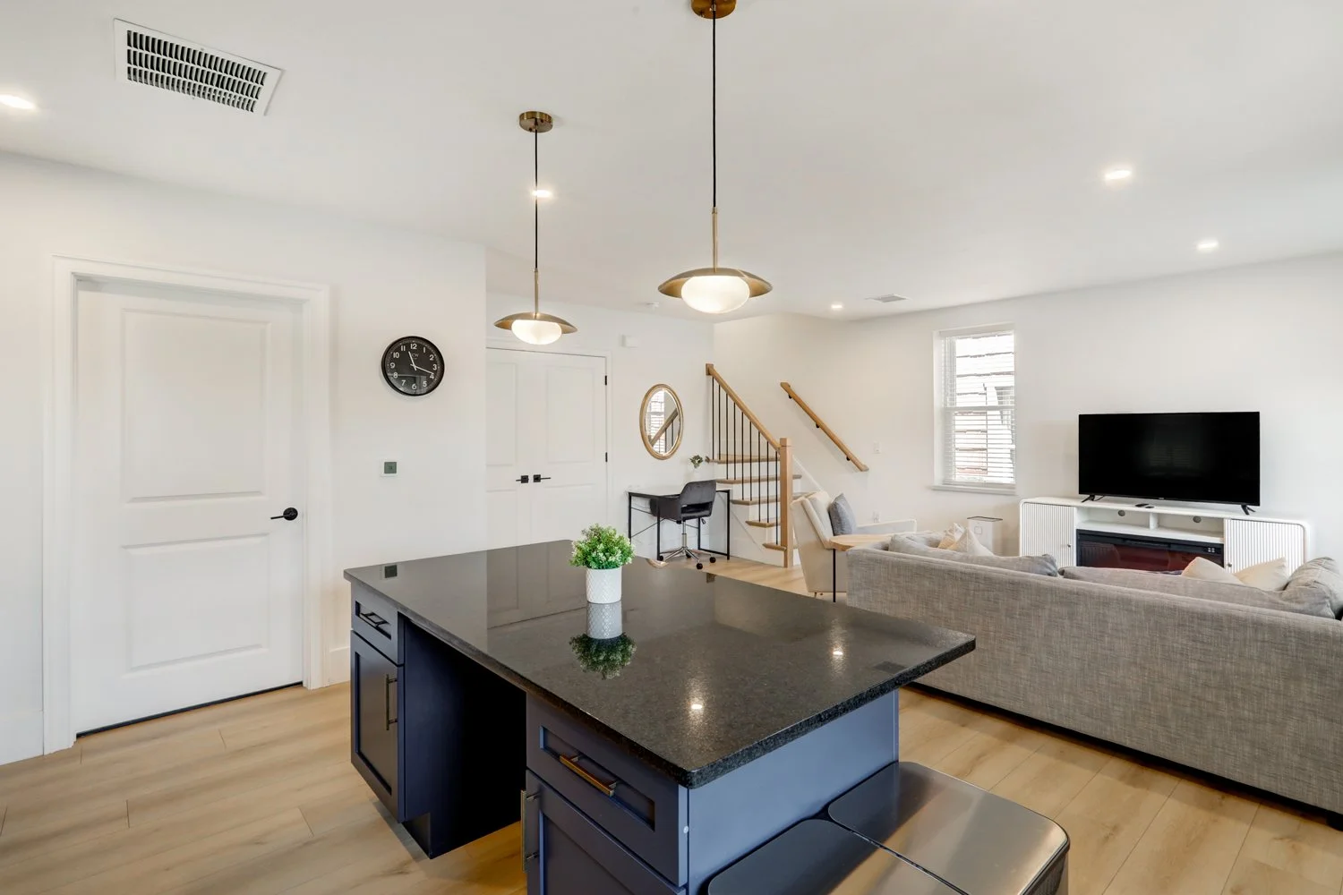 Open-plan living room and kitchen with white walls, hardwood floors, a black kitchen island, gray sofas, a TV on a white console, a small desk with a mirror, and a staircase with wooden steps and black railing.