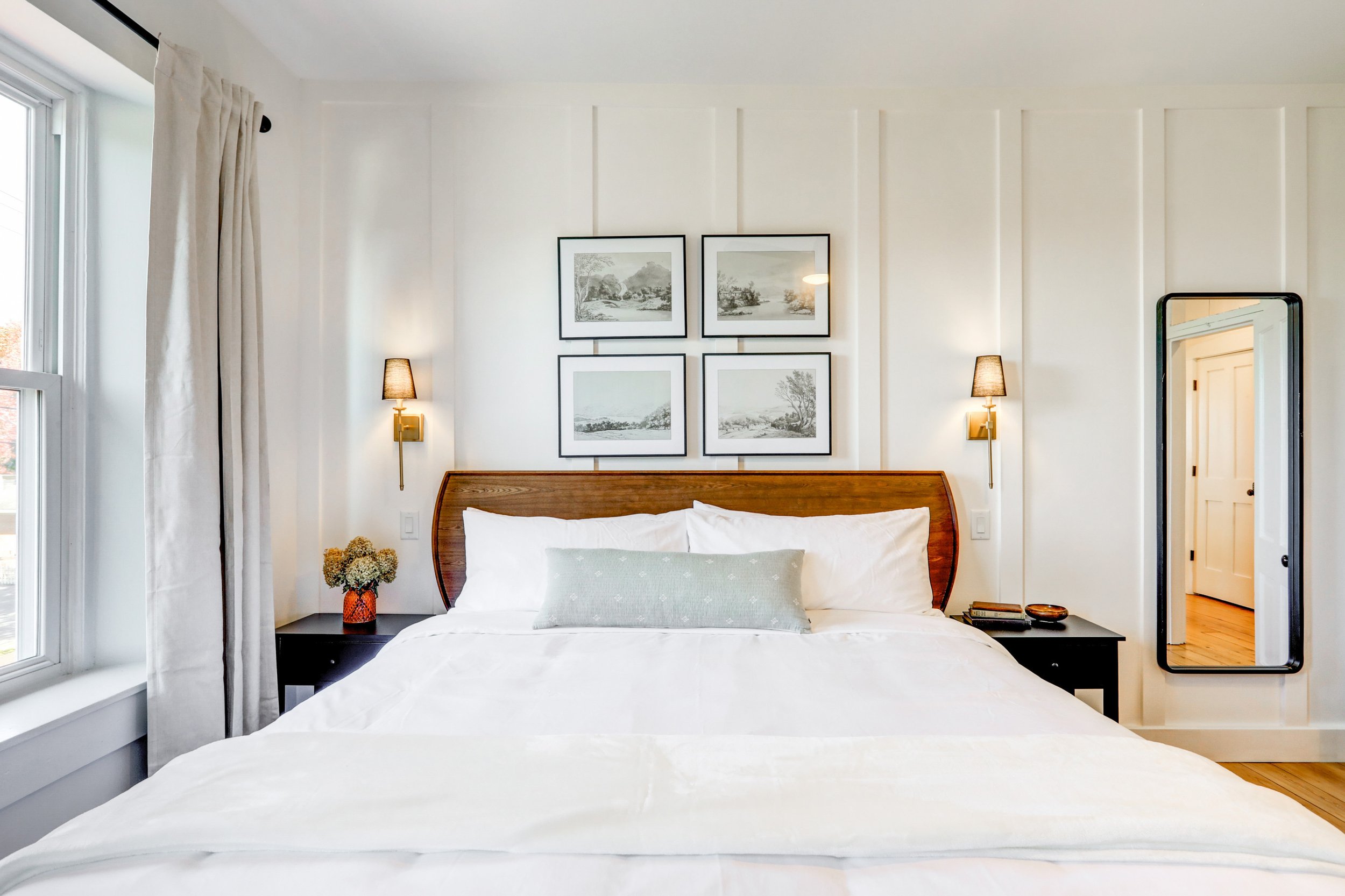 A cozy bedroom with a large bed, white bedding, a wooden headboard, and a decorative pillow. Two black bedside tables with matching lamps, framed black-and-white landscape photos on the white wall above the bed, a vase with flowers, and a full-length