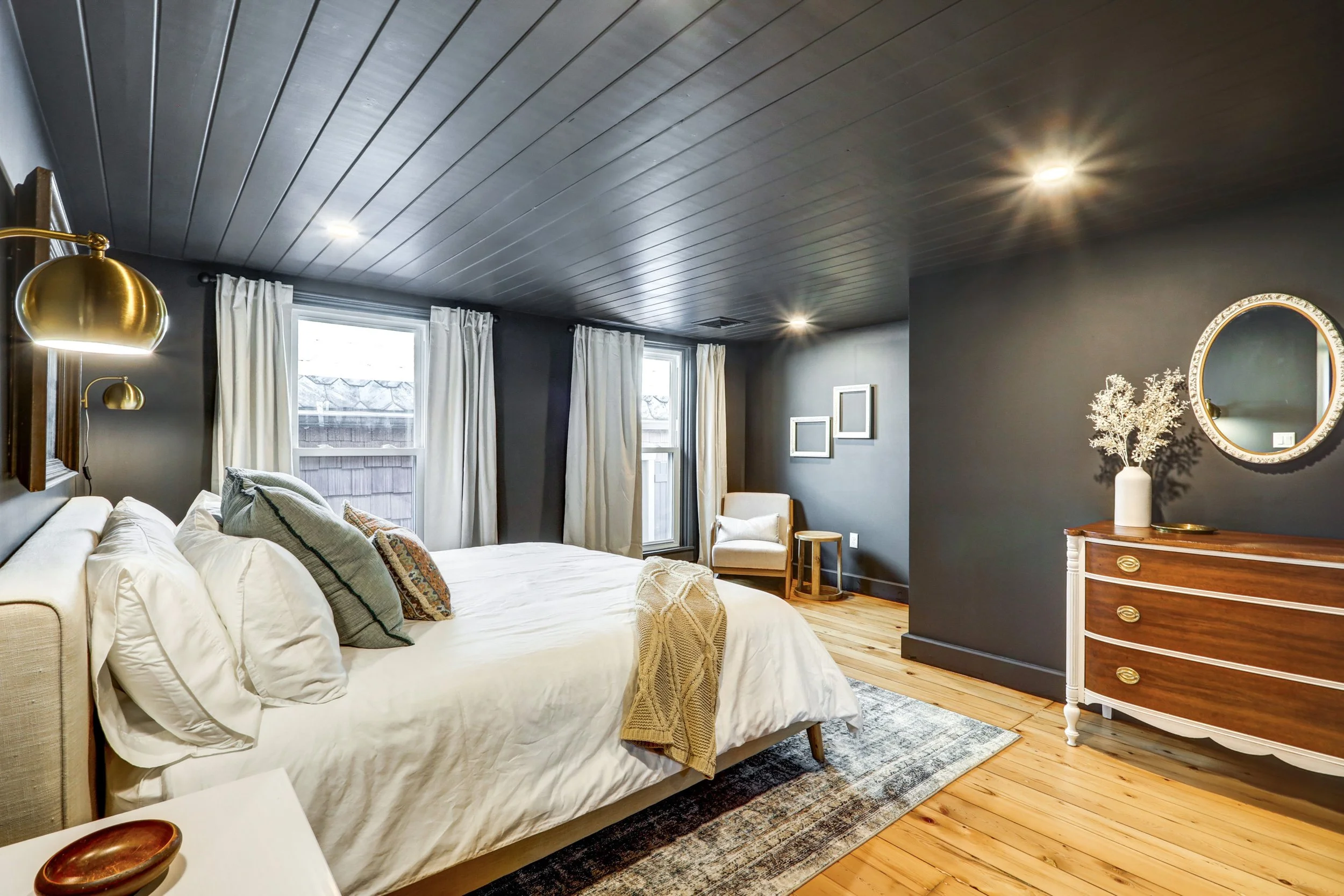 A bedroom with a white bed, beige pillows, wooden floor, gray walls, two windows with white curtains, a dresser with a mirror, a vase with dried flowers, and brass wall-mounted lamps.