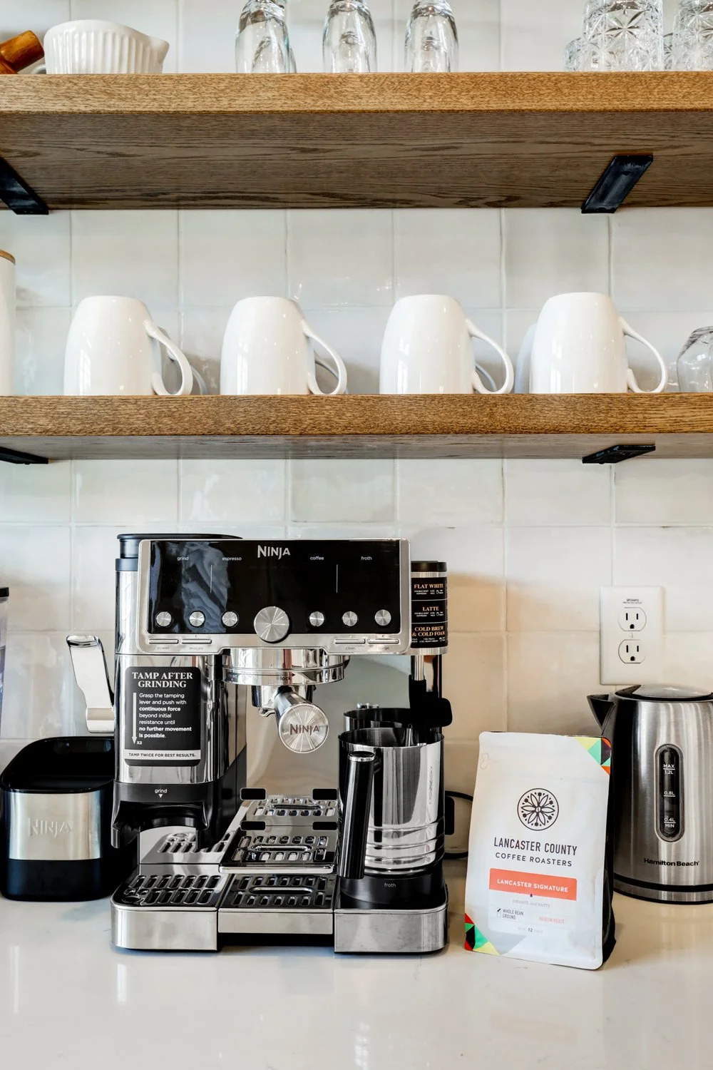 A coffee station with a Ninja espresso machine, a bag of Lancaster County coffee, a stainless steel kettle, and four white mugs on a shelf, against a tiled wall.