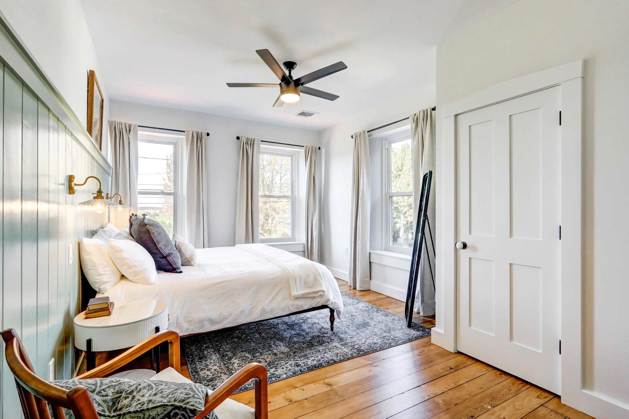 Bright bedroom with white walls, large windows with white curtains, a bed with white bedding and pillows, dark area rug, wooden floors, and ceiling fan.