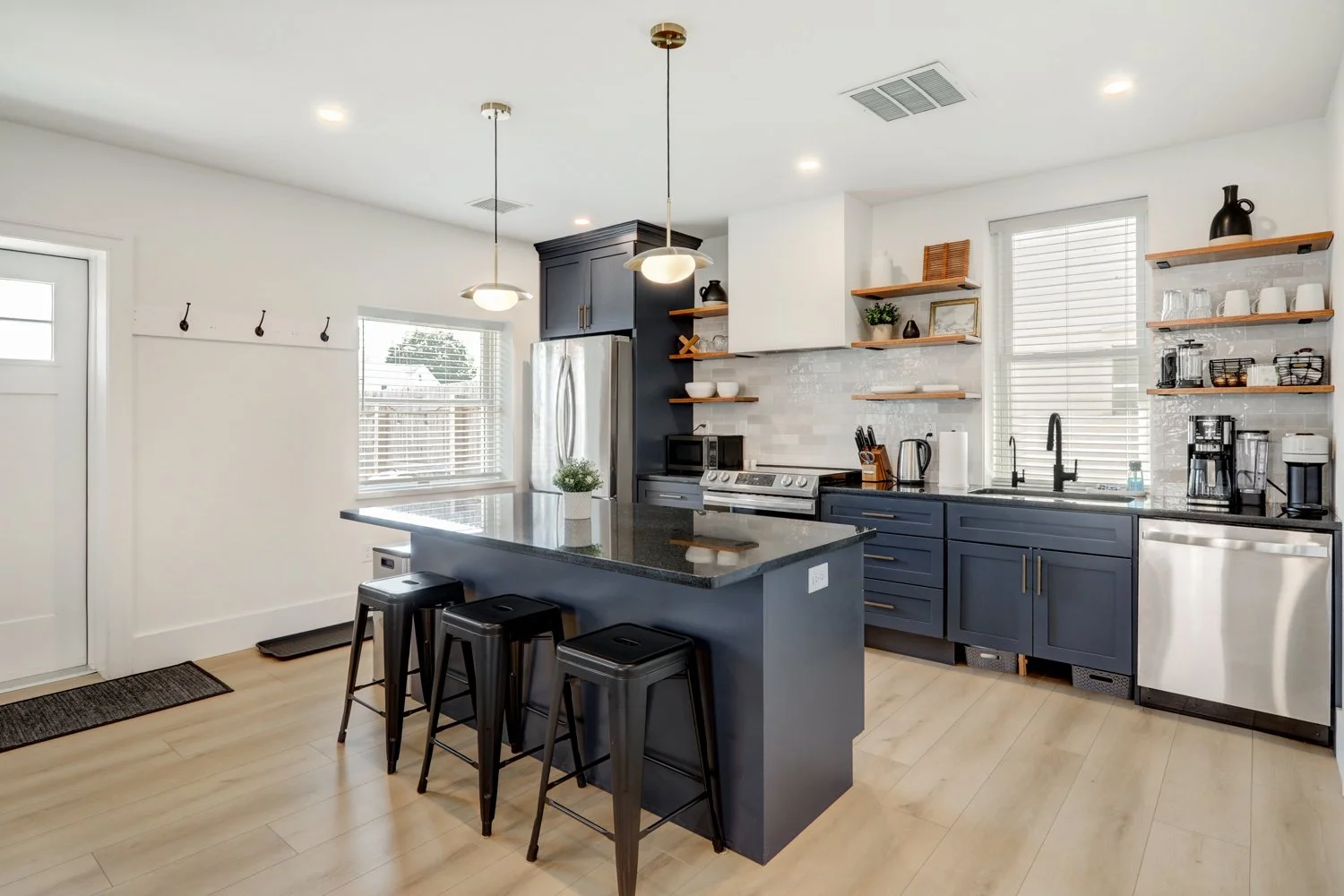 Modern kitchen with navy cabinets, wooden open shelves, stainless steel appliances, black countertop island with bar stools, light wood flooring, and white walls with large windows.