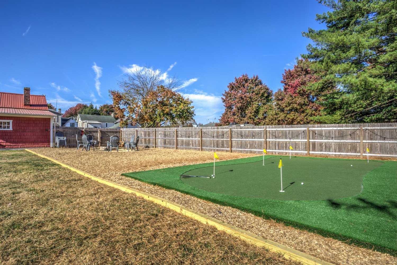 Backyard with a putting green, a fenced area with leaf-like mulch, several chairs near the fence, and trees with fall foliage against a blue sky.