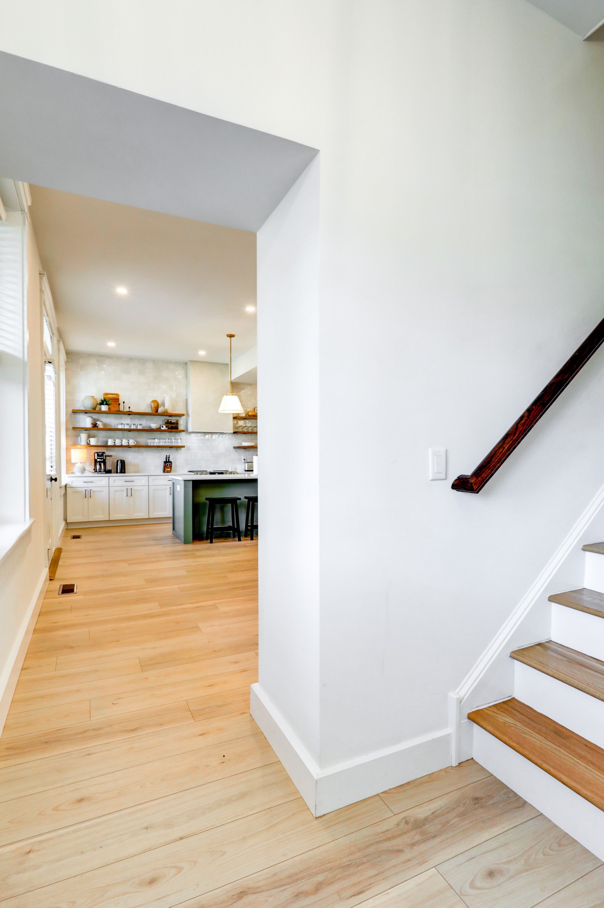Interior view of a modern kitchen with white cabinets, open wooden shelves with dishes and decor, a small kitchen island with stools, light-colored wooden flooring, and a staircase with wooden steps and a dark wood railing.