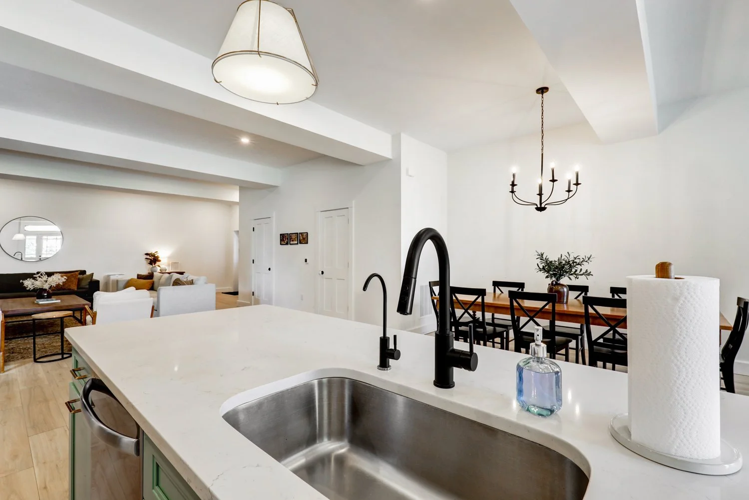Open-concept living and dining area with white walls, wooden flooring, a kitchen island with a sink, and a dining table with black chairs. Modern lighting fixtures include a chandelier and a pendant lamp.