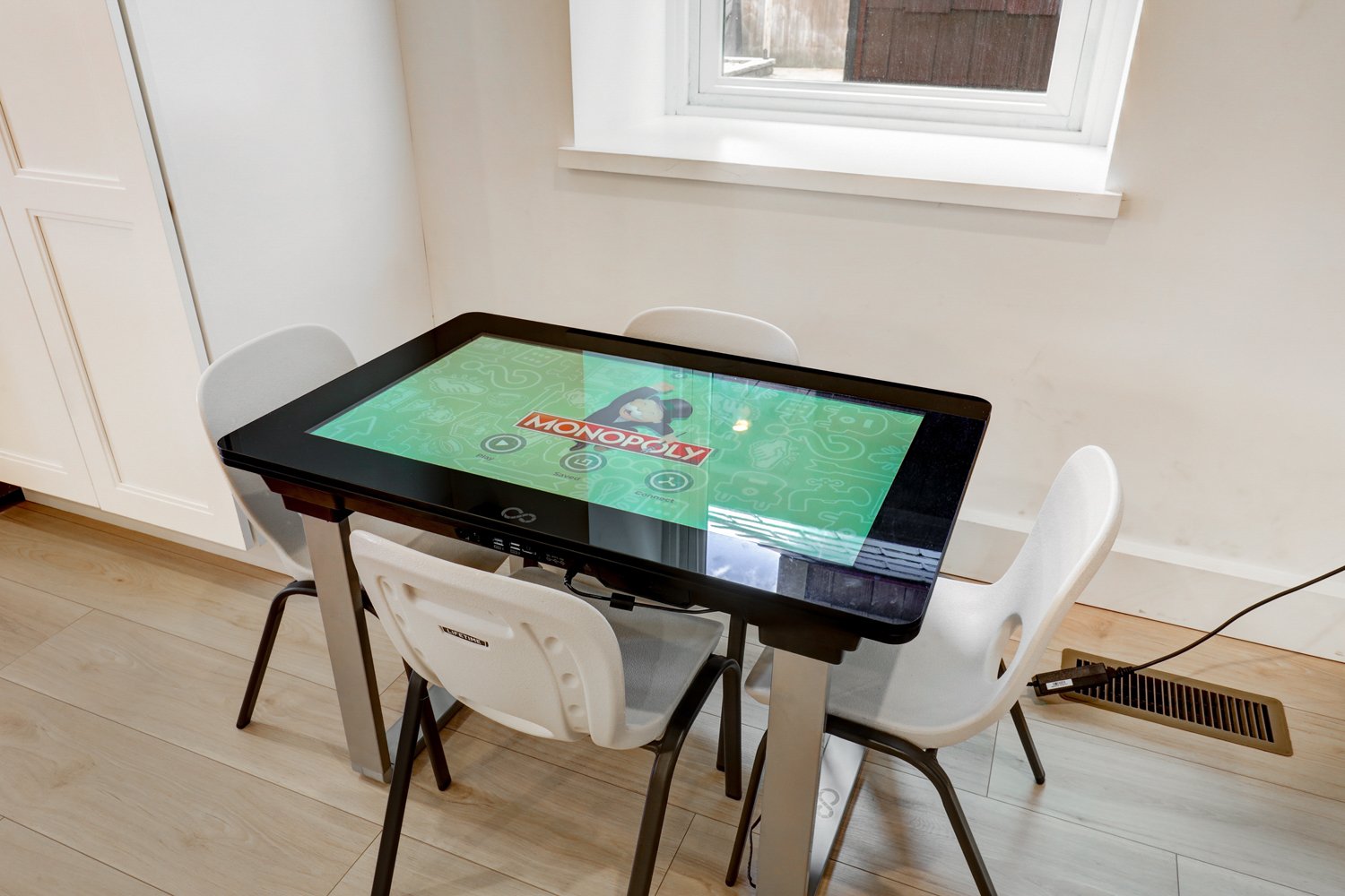 A kid's table with a digital touchscreen playing Monopoly Junior, surrounded by three white chairs, in a bright room with wooden flooring, a window, and an air vent.