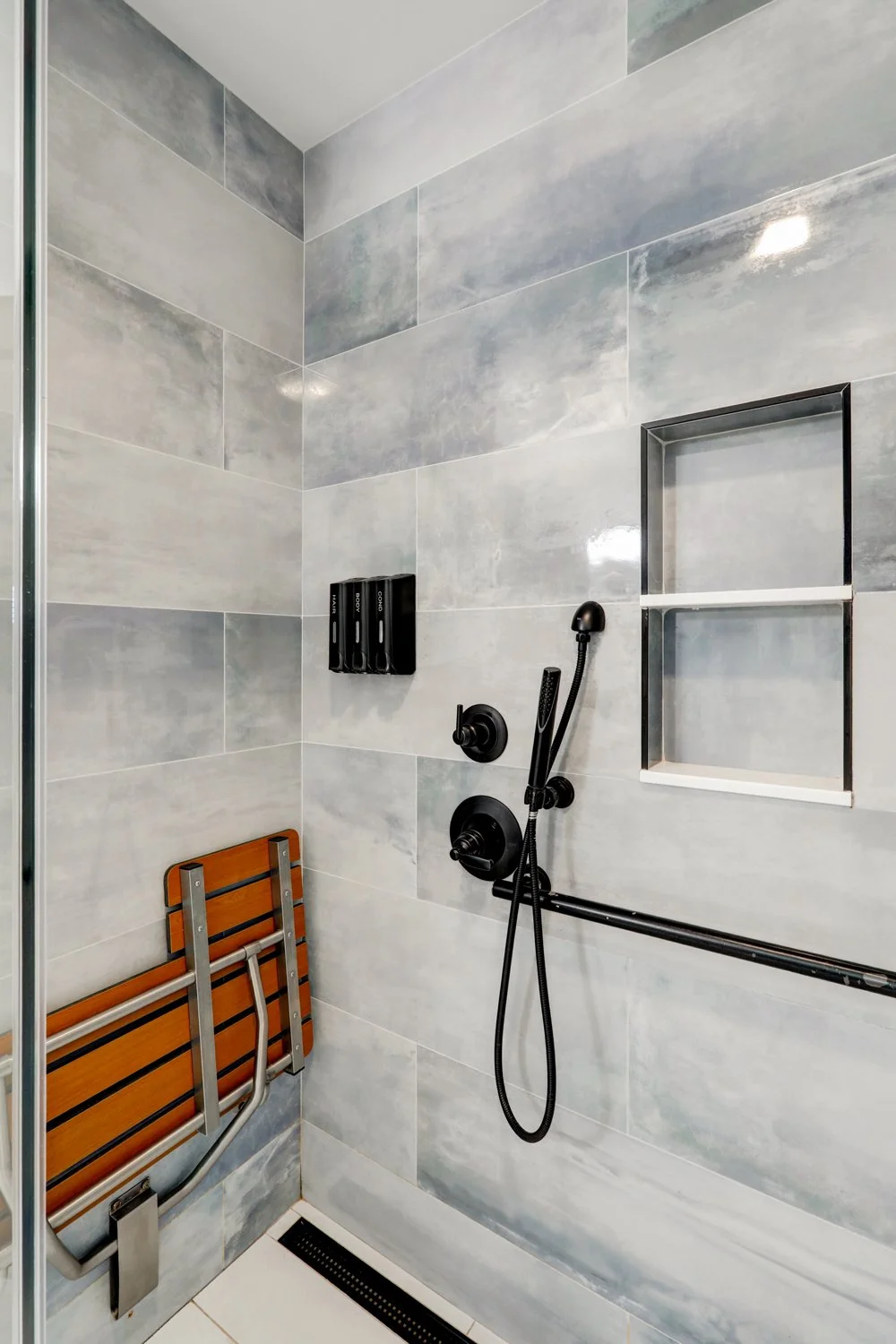 Shower with gray marble tiles, black fixtures, a wooden fold-down shower seat, built-in shelf, and a grab bar