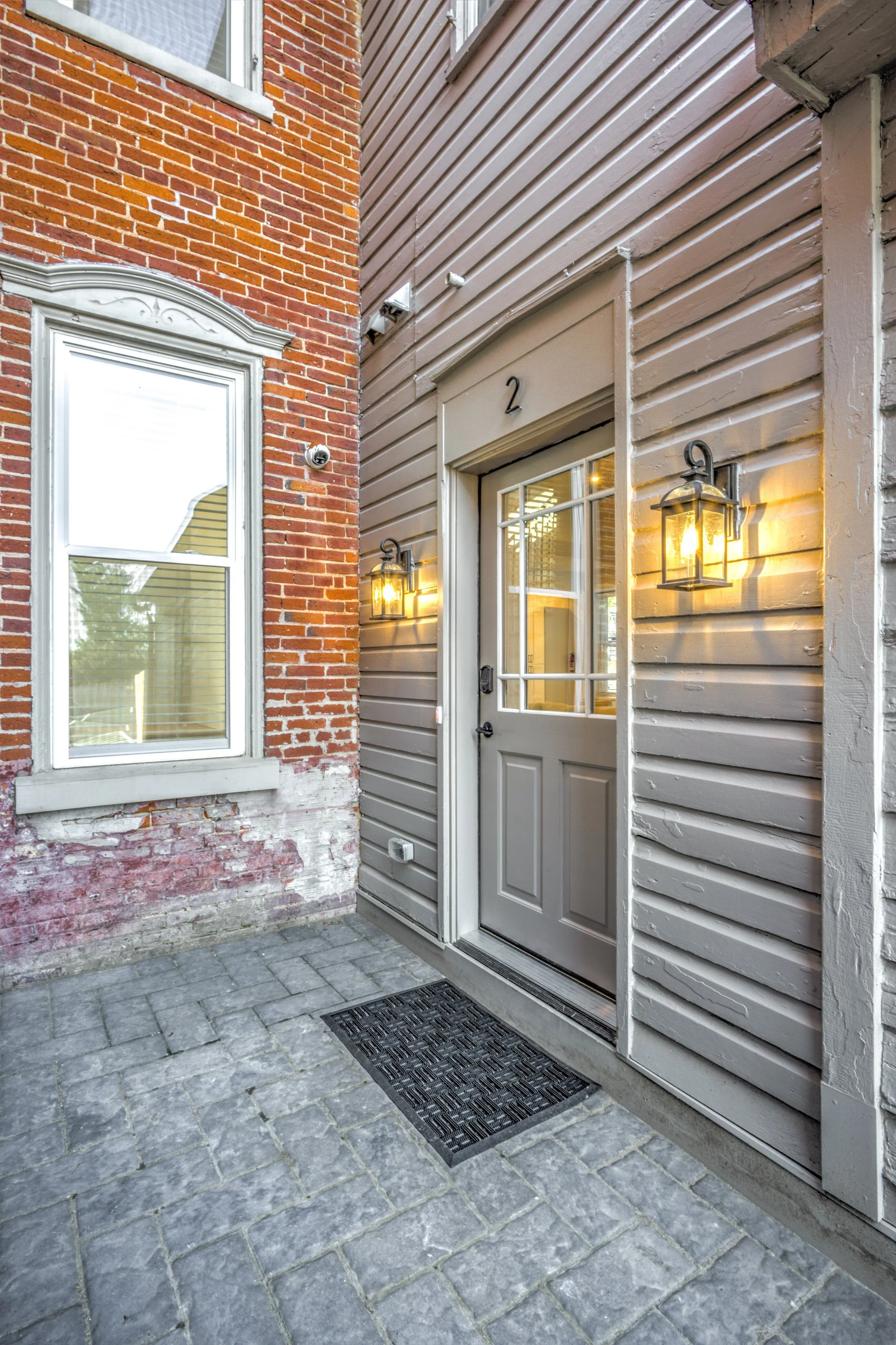 The image shows a residential entryway with a gray door and a glass window, flanked by two vintage-style wall lanterns with warm lighting. The door is under a beige-colored, horizontally paneled siding, with a house number '2' above it. To the left i