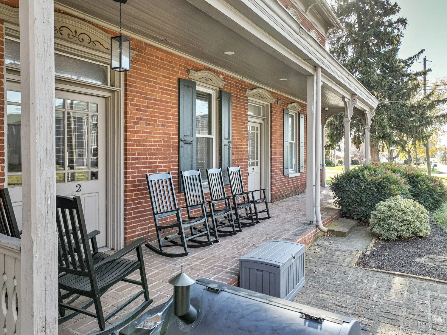 Front porch with four black rocking chairs, brick pathway, and green bushes in front of a red brick building with blue shutters and white trim.