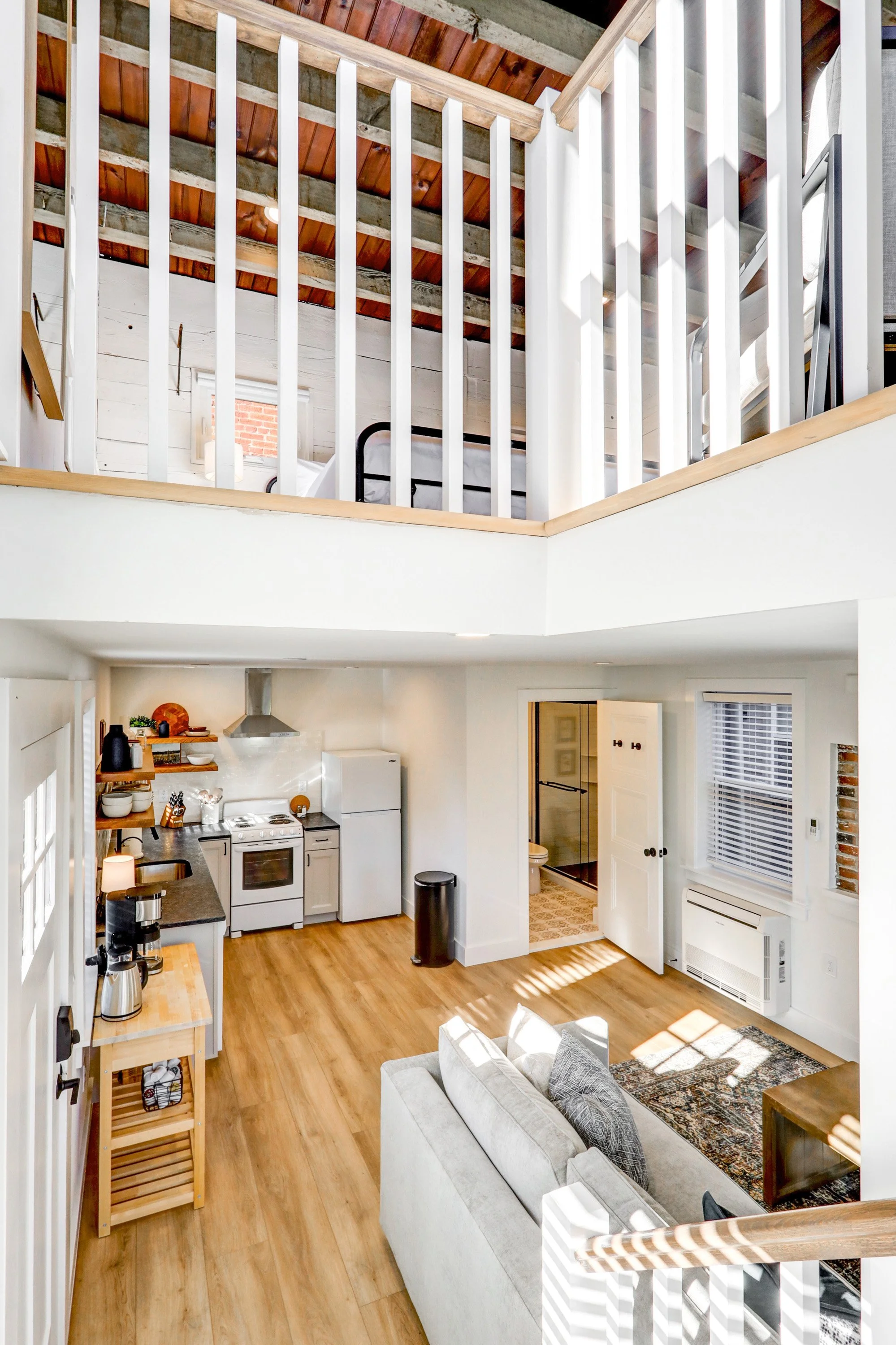 Interior view of a cozy, modern living space with a kitchen and a living room. The kitchen features white appliances, wooden shelves, and a small wooden serving cart. The living room has a light-colored sofa with patterned pillows and a vintage-style